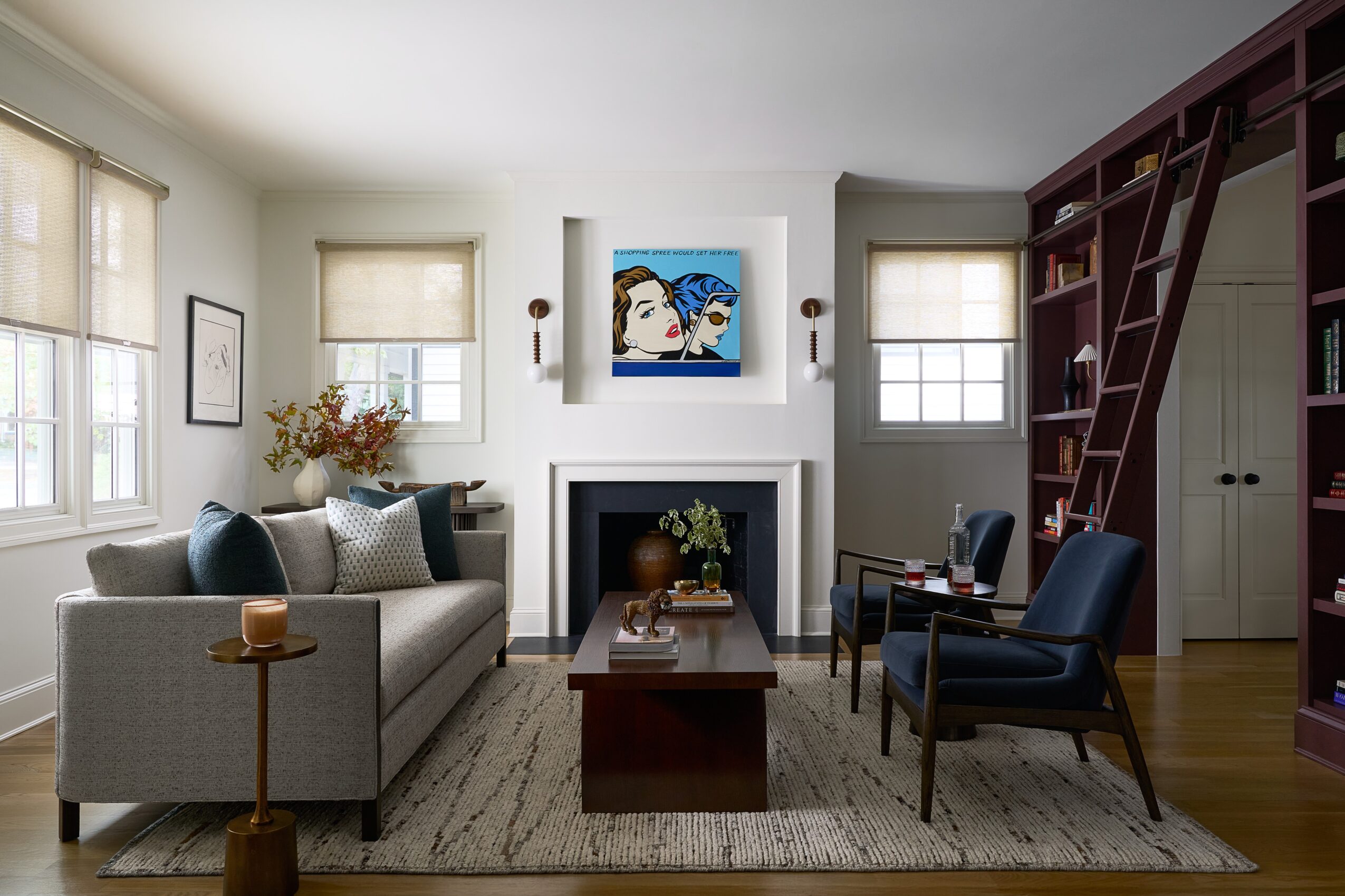 Living room with sofa, armchair, fireplace, bookshelf, and artwork, illuminated by natural light from windows.