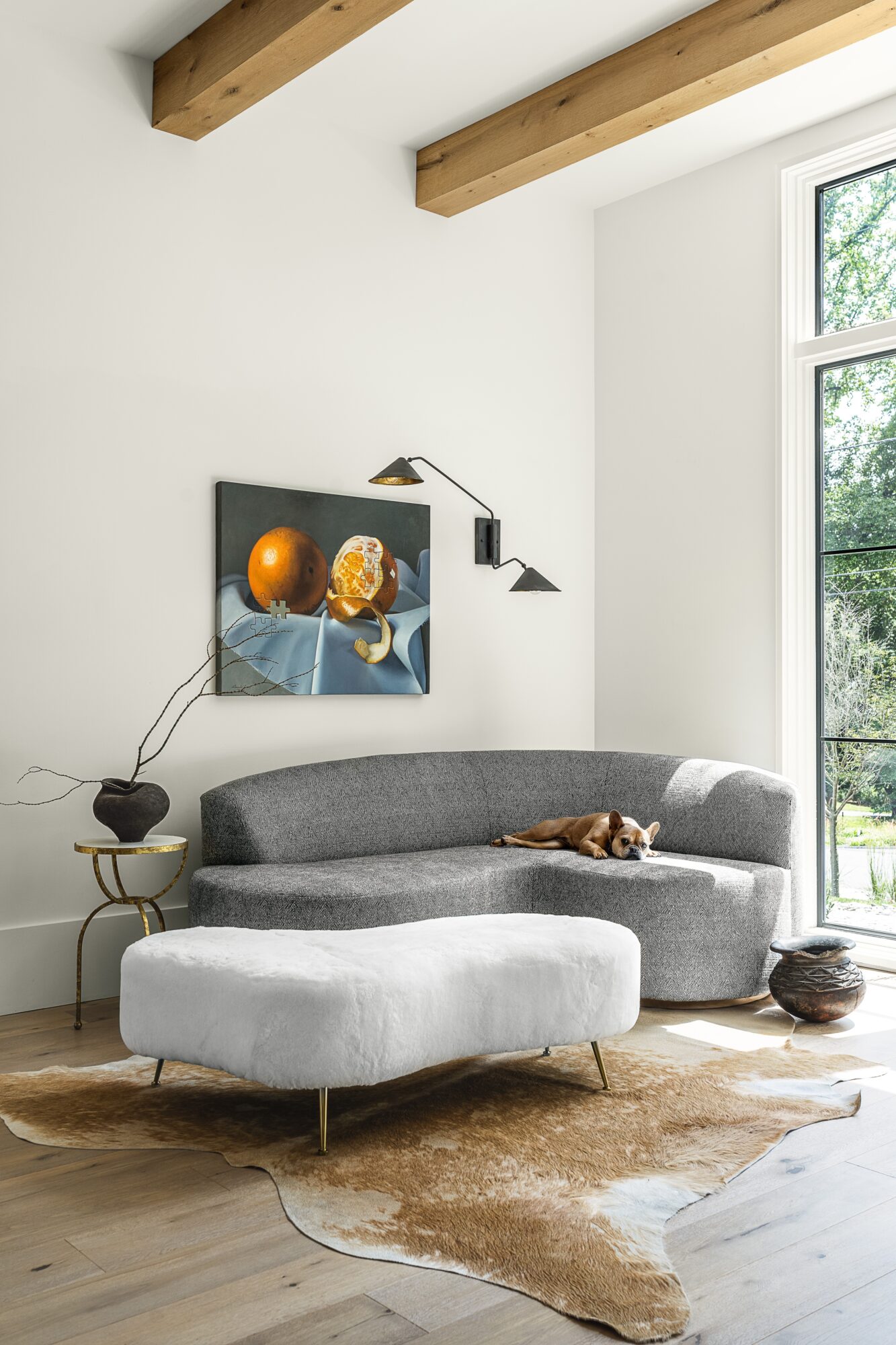 Living room with a gray sectional sofa, a white ottoman, a wall-mounted lamp, and a window with trees outside.