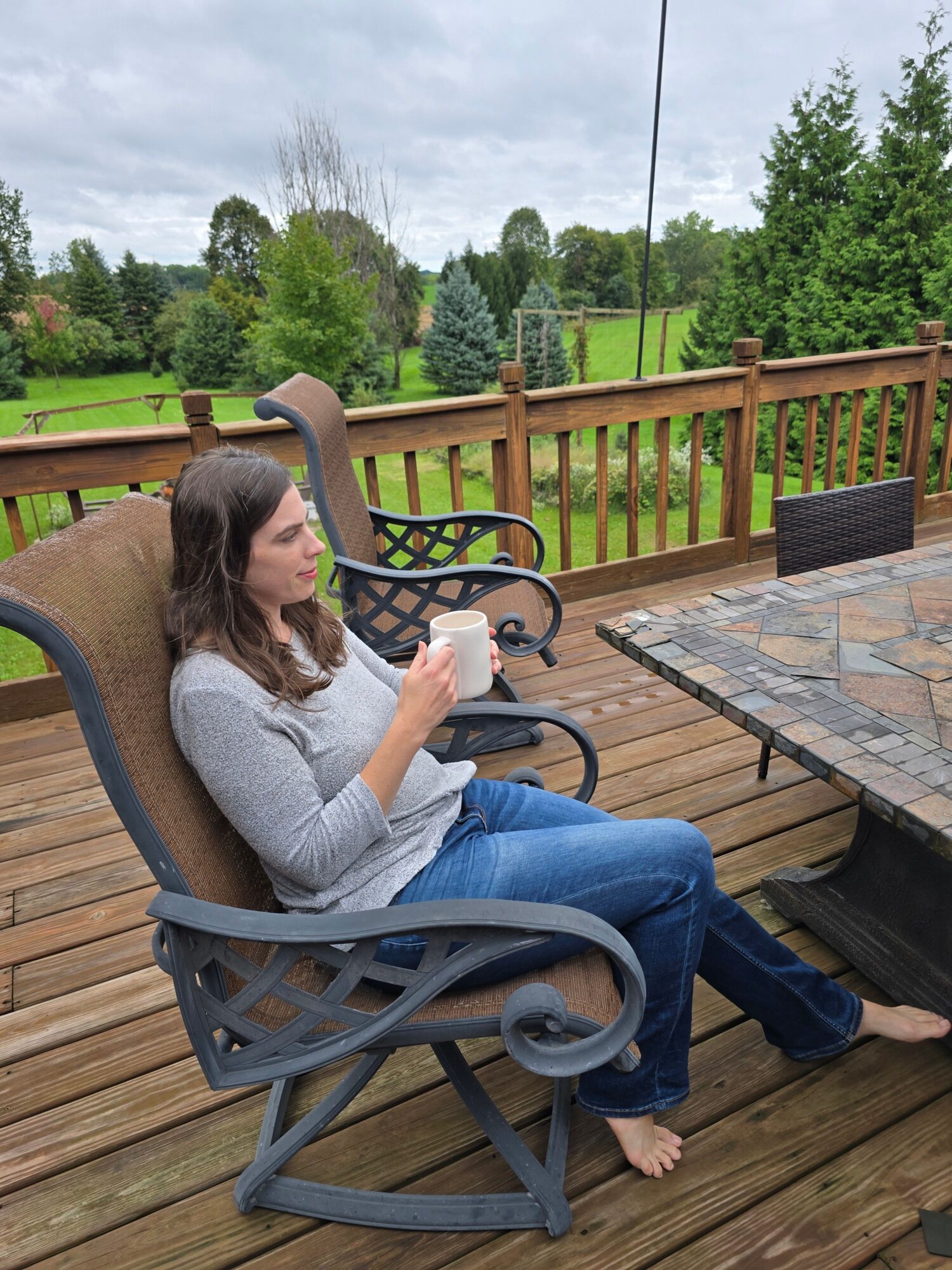 Woman sitting on a wooden deck, holding a cup, with a garden and trees in the background.