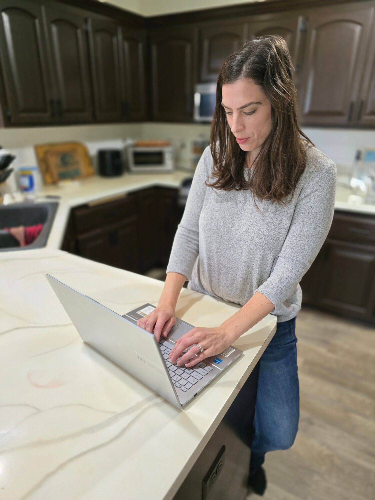 Woman typing on a laptop in a kitchen with dark cabinets and a white countertop.