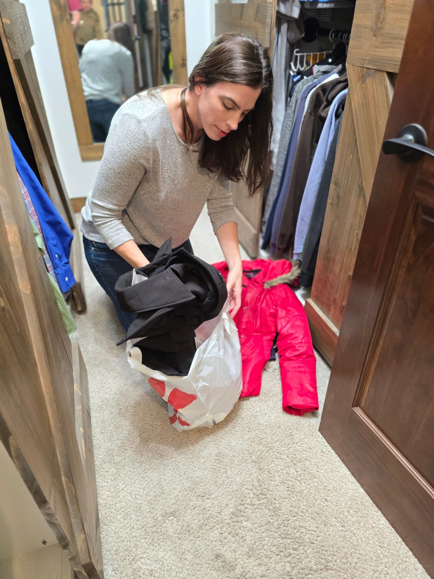 Woman with brown hair, wearing a beige sweater, holding a black bag and a white plastic bag, standing in a room with a closet.