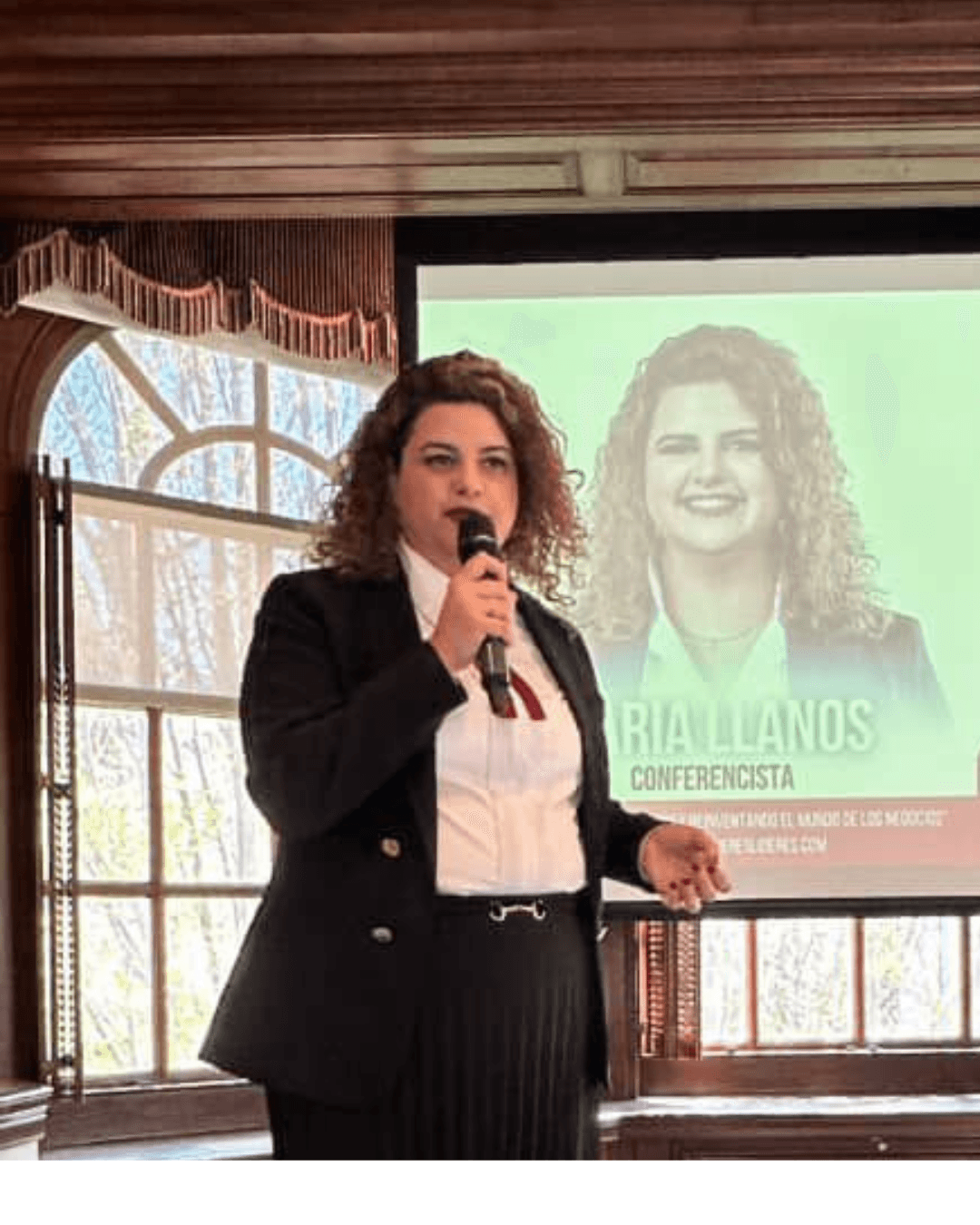 Woman speaking into microphone during presentation, with a large screen showing her image behind her, in a room with windows.