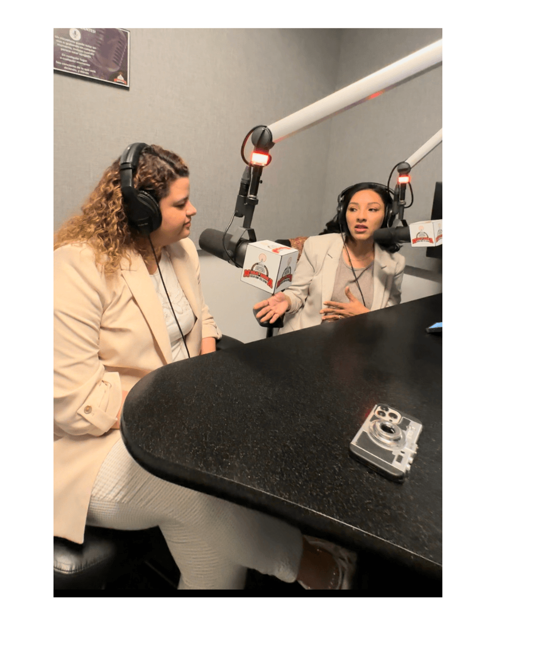 Two women sit at a table with microphones and headphones, engaged in conversation in a radio studio.