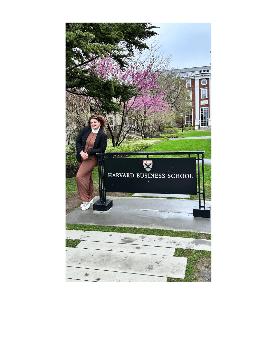 Woman standing next to Harvard Business School sign with trees and a building in background.