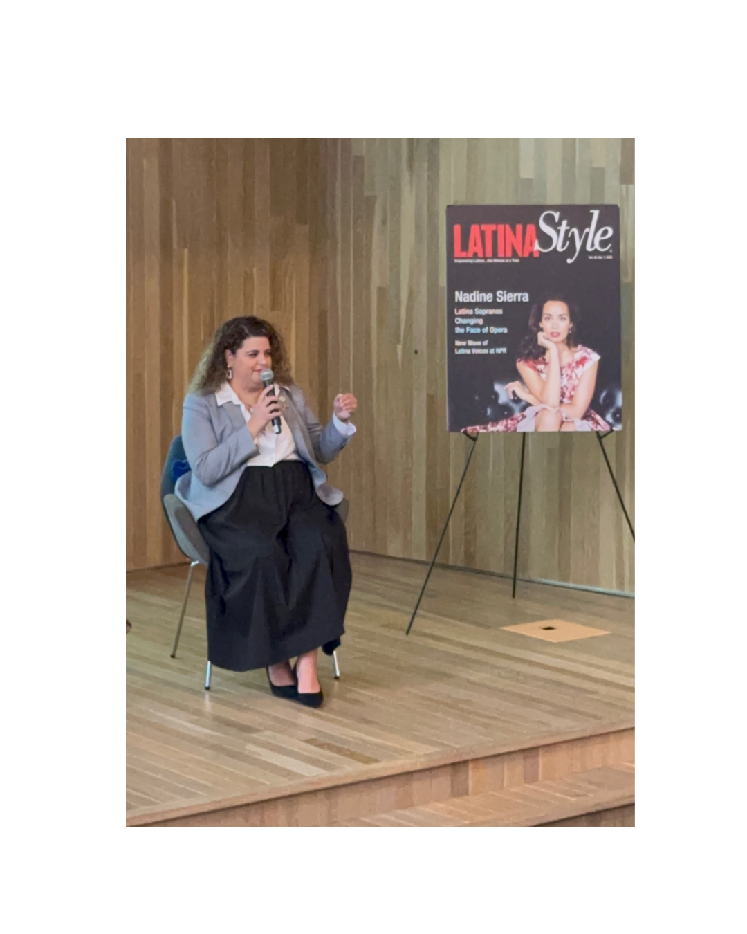 Woman sitting on a chair speaking into a microphone, next to a poster on an easel, in a room with wooden walls and floor.