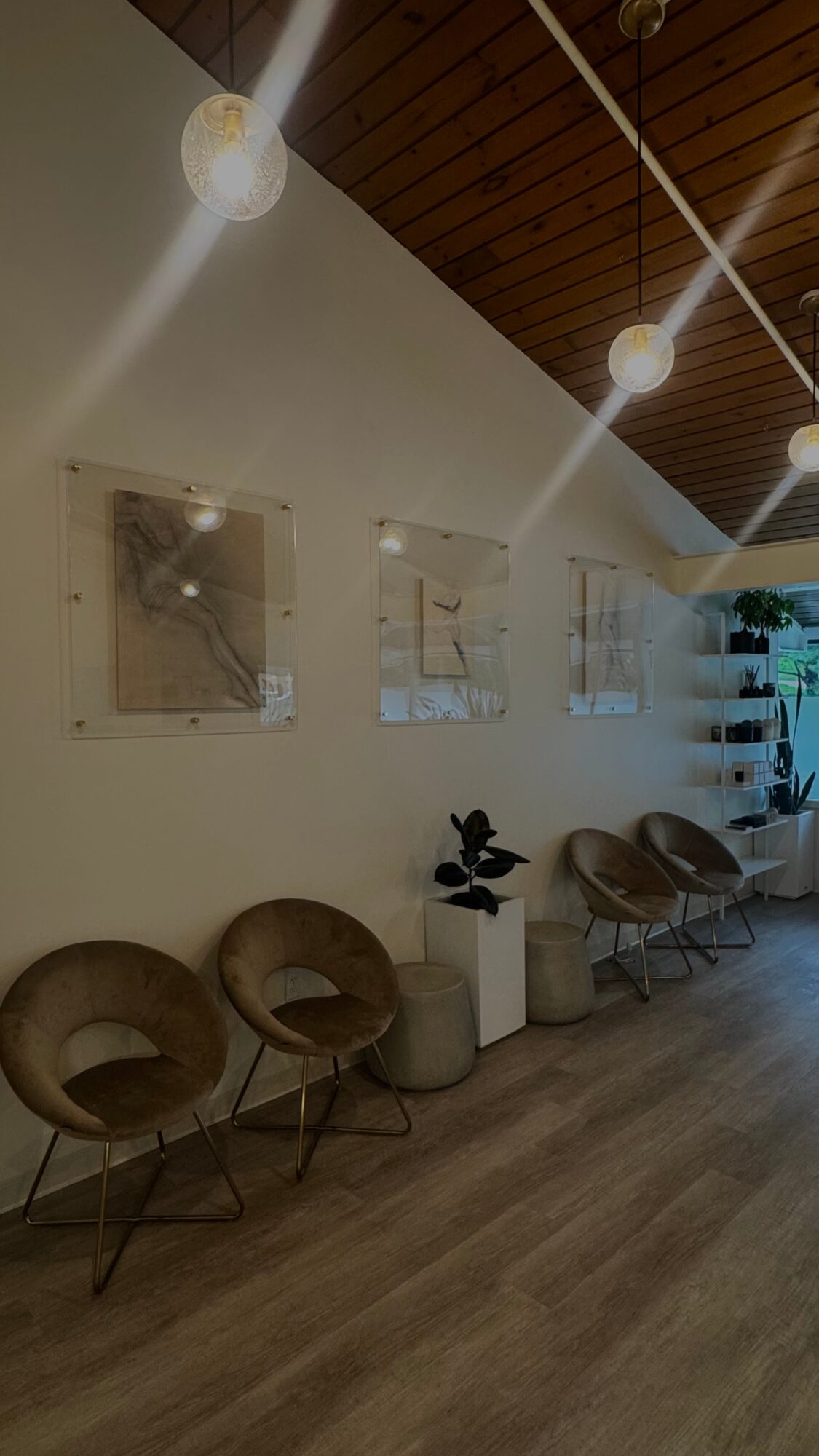 Interior space with three brown chairs, white wall with artwork, and ceiling lights, wooden ceiling, and floor.
