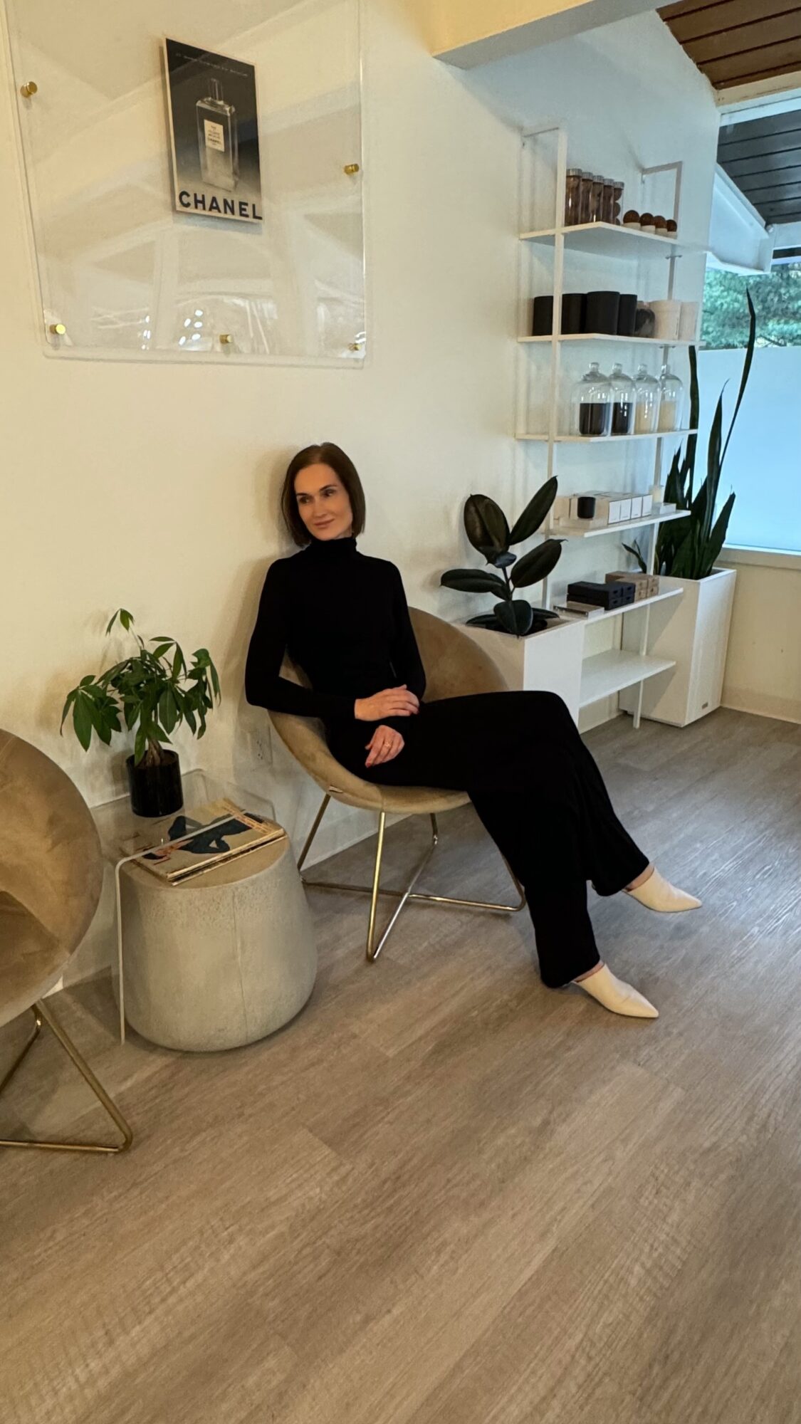 Woman in black sitting on a chair in a room with white shelves and plants, wooden floor, and a window.