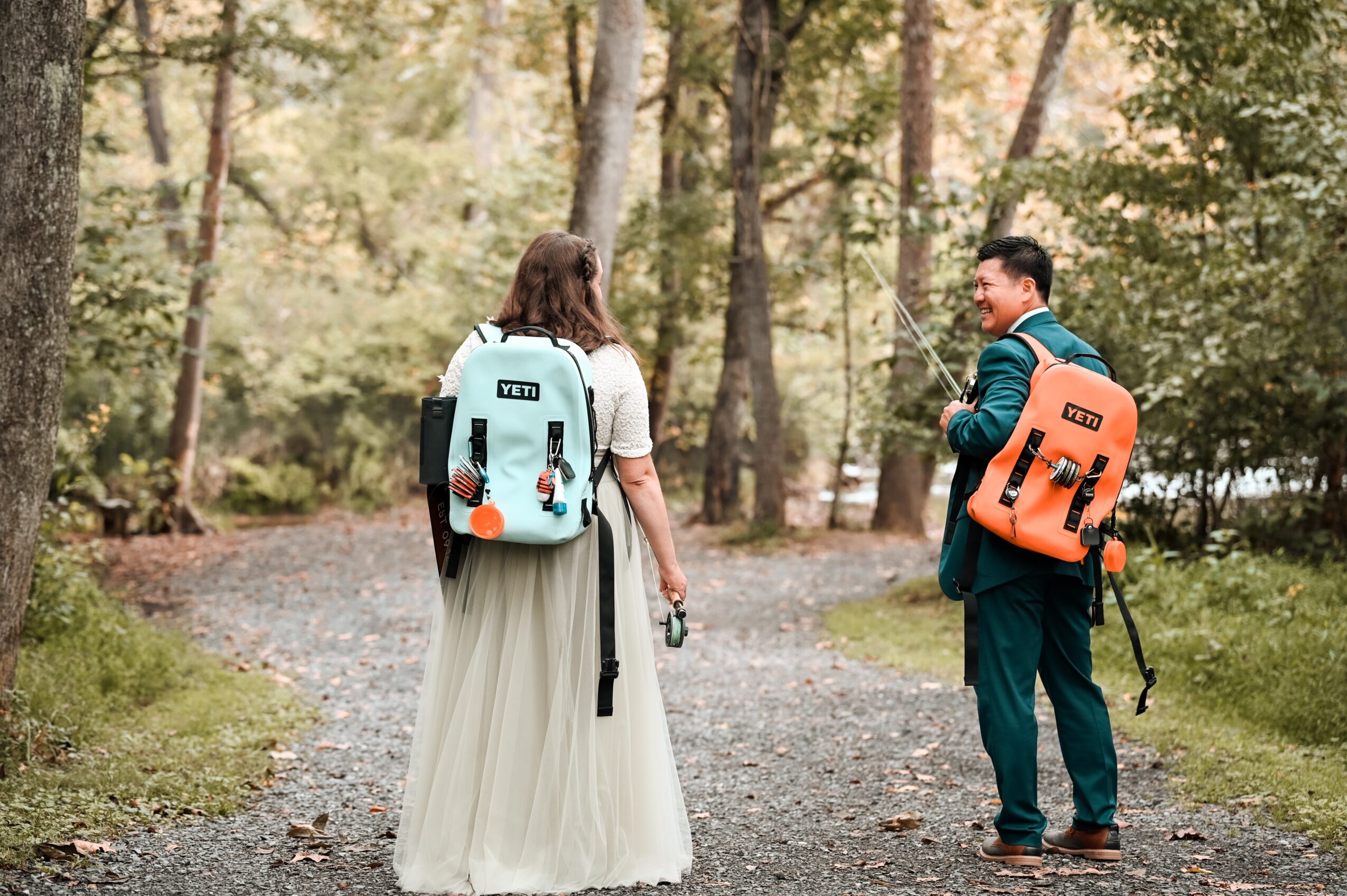 Two people with backpacks walking on a forest path, trees in the background, one in a long skirt and the other in a suit.