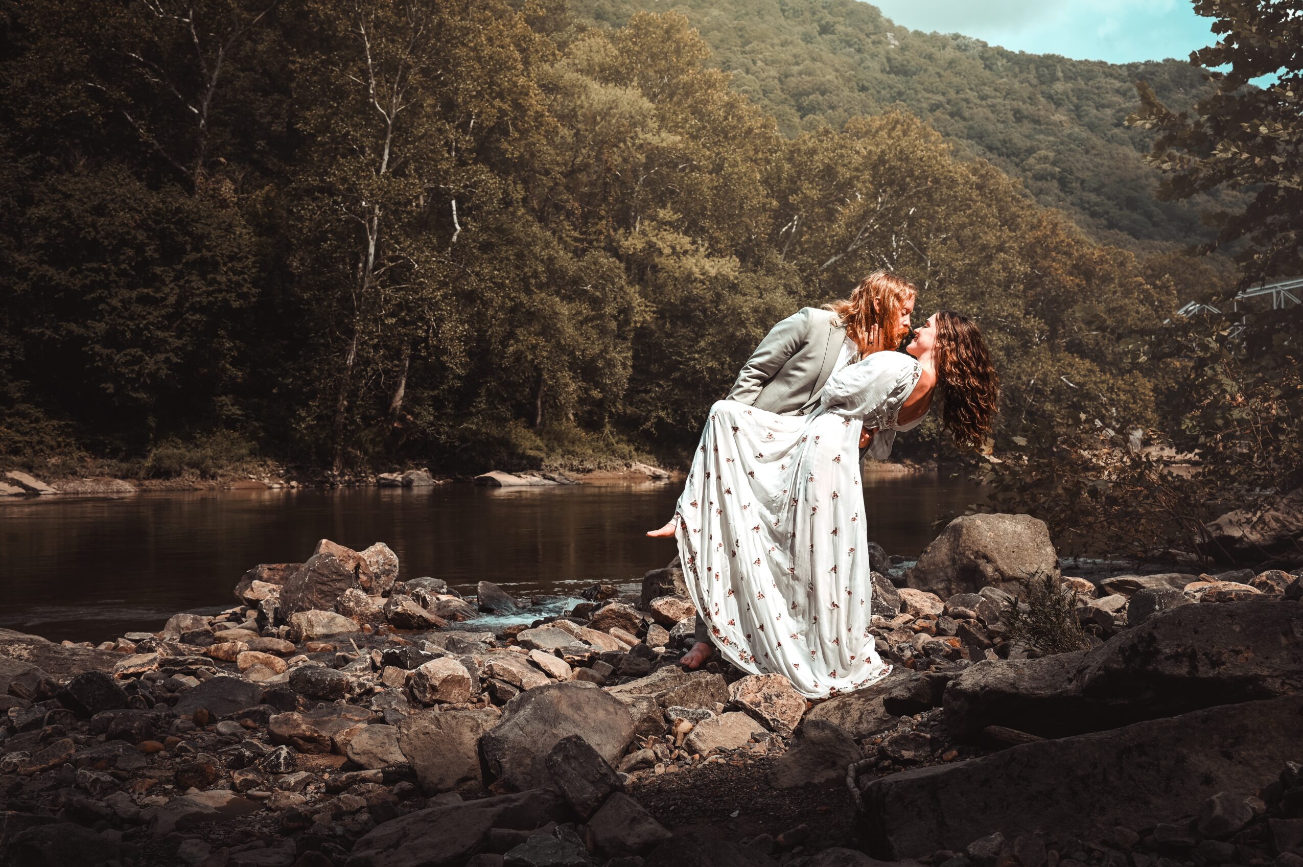 Two people, one with long red hair, are near a riverbank surrounded by trees, with one leaning over the other.