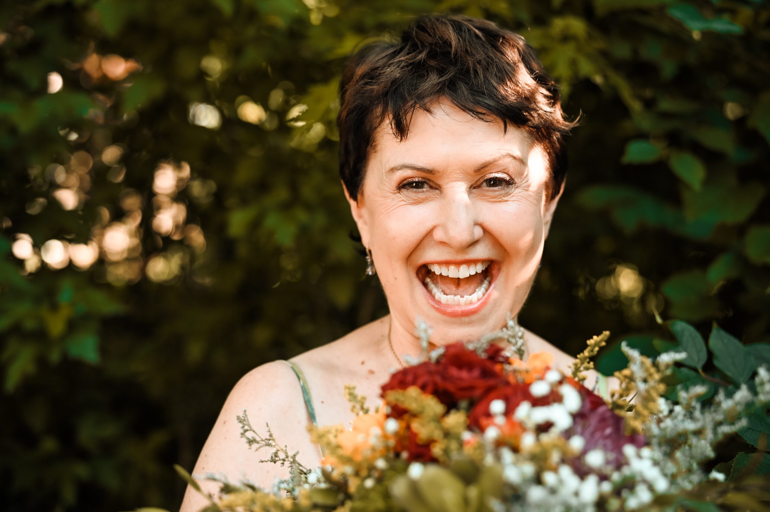 Woman with short dark hair smiling outdoors holding a bouquet of flowers, surrounded by greenery.