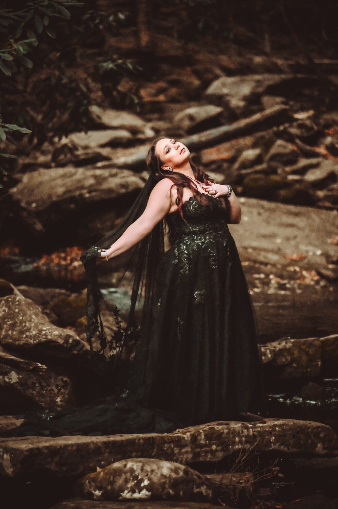 Woman in a black dress standing on rocks outdoors, head tilted back, arms raised, with a natural rocky background.