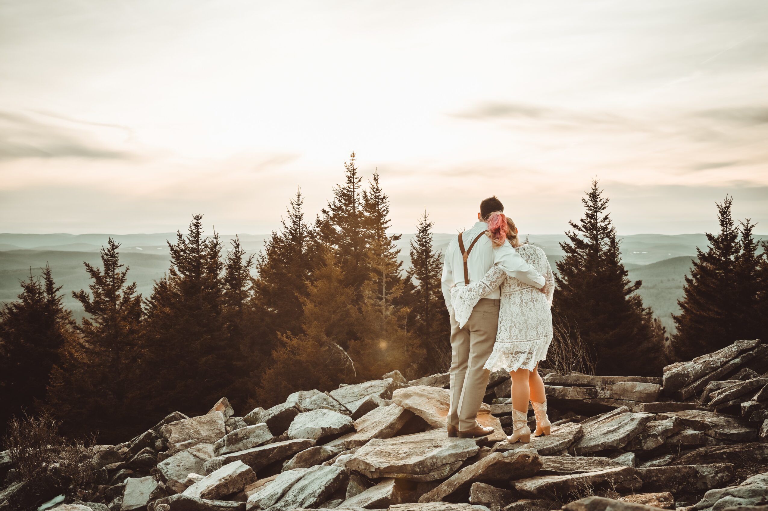 A couple hugging on rocky terrain with trees and a cloudy sky in the background.