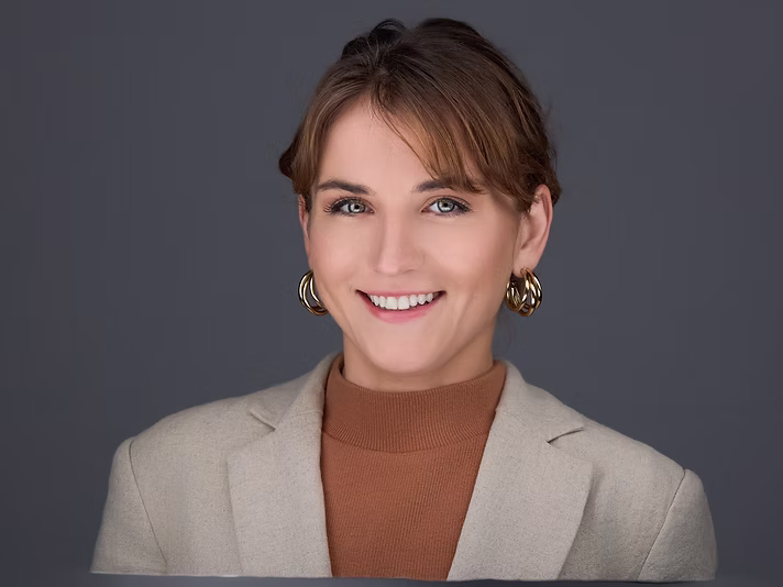 Portrait of a smiling woman with short brown hair, wearing a beige blazer and gold hoop earrings, against a gray background.