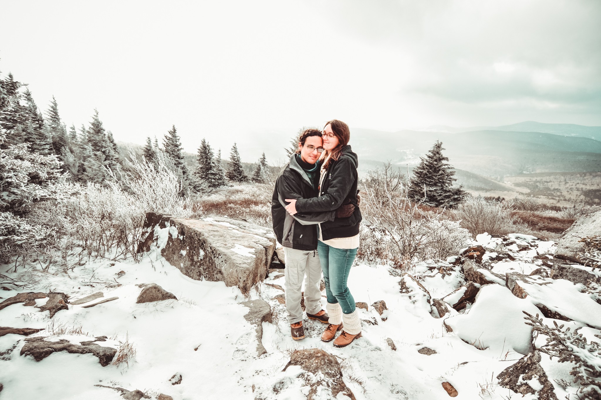 Two people embrace on snow-covered rocks with a winter landscape and distant mountains in the background.