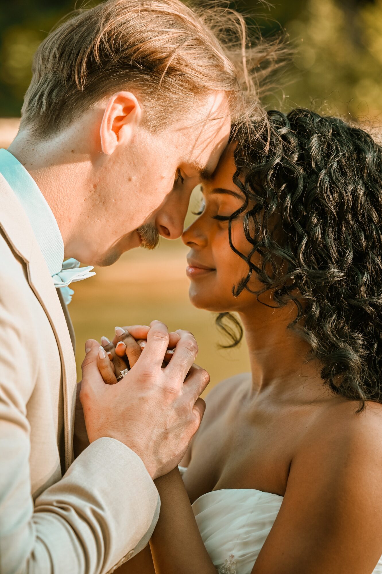 A couple with foreheads touching, eyes closed, holding hands outdoors during daytime, smiling softly.