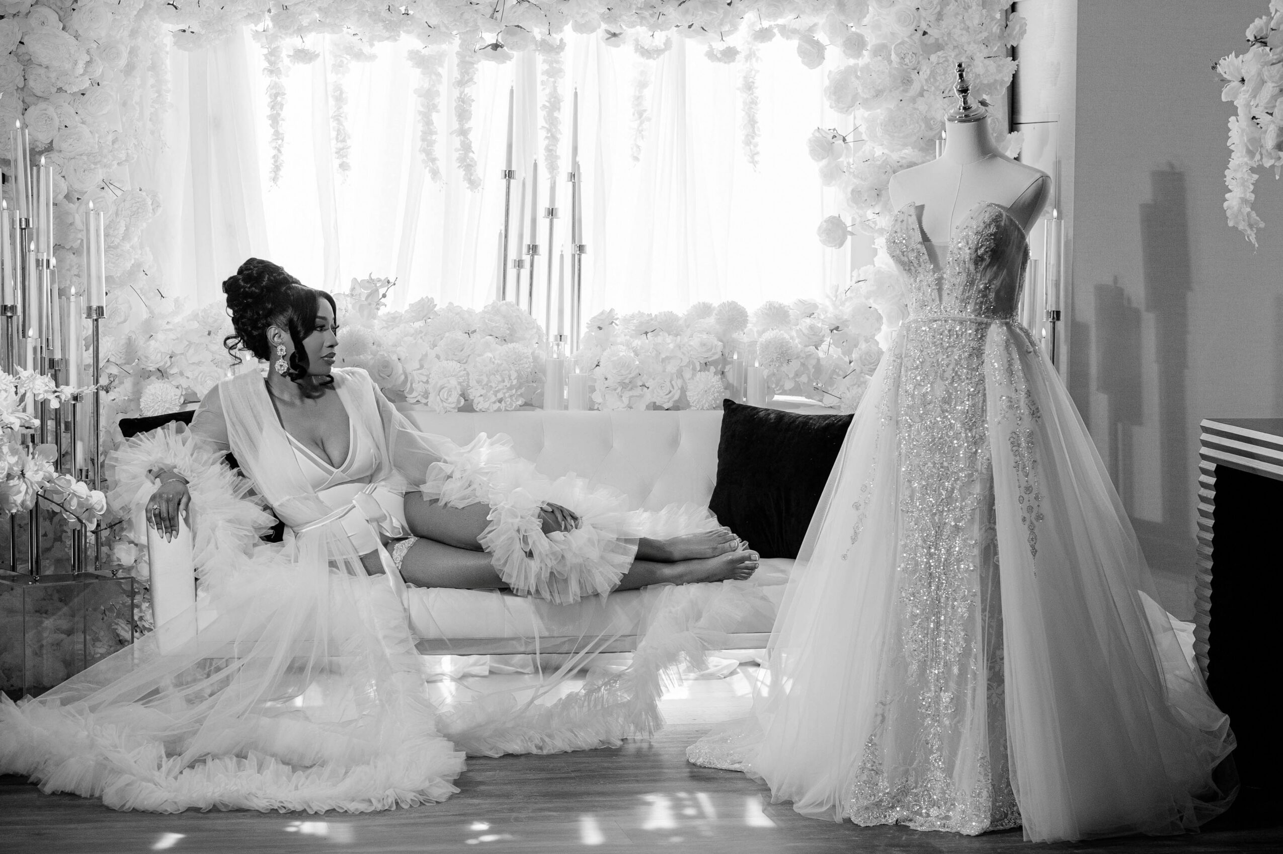 Woman in wedding dress sitting on sofa, looking at a wedding gown on a mannequin, in a bright room with curtains.