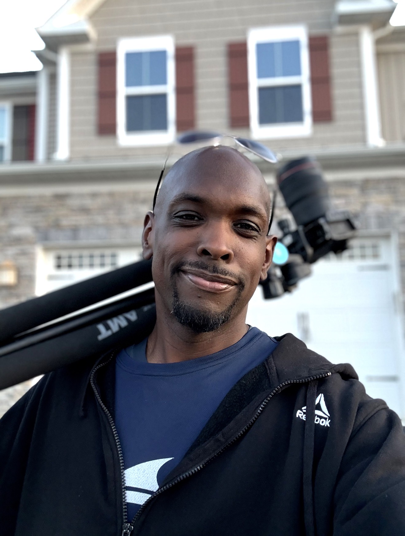 Man taking a selfie outdoors in front of a house, holding a camera tripod on his shoulder, smiling.