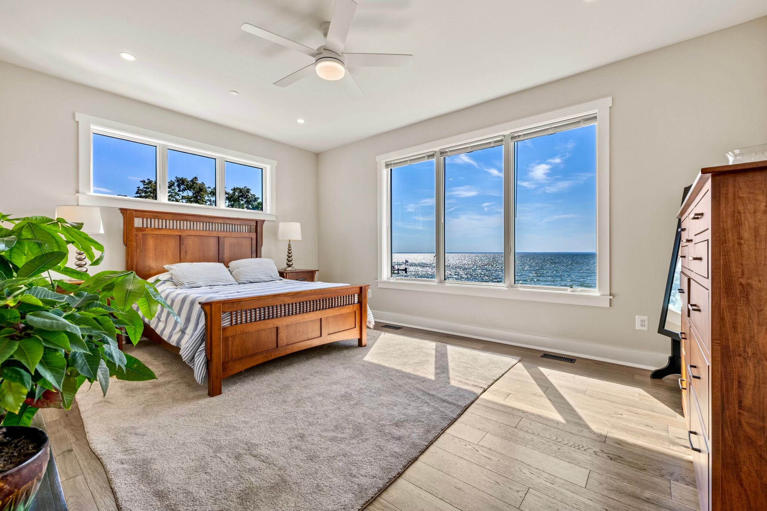 Bedroom with large windows showing a view of the sky and water, wooden bed, potted plant, dresser, and ceiling fan.