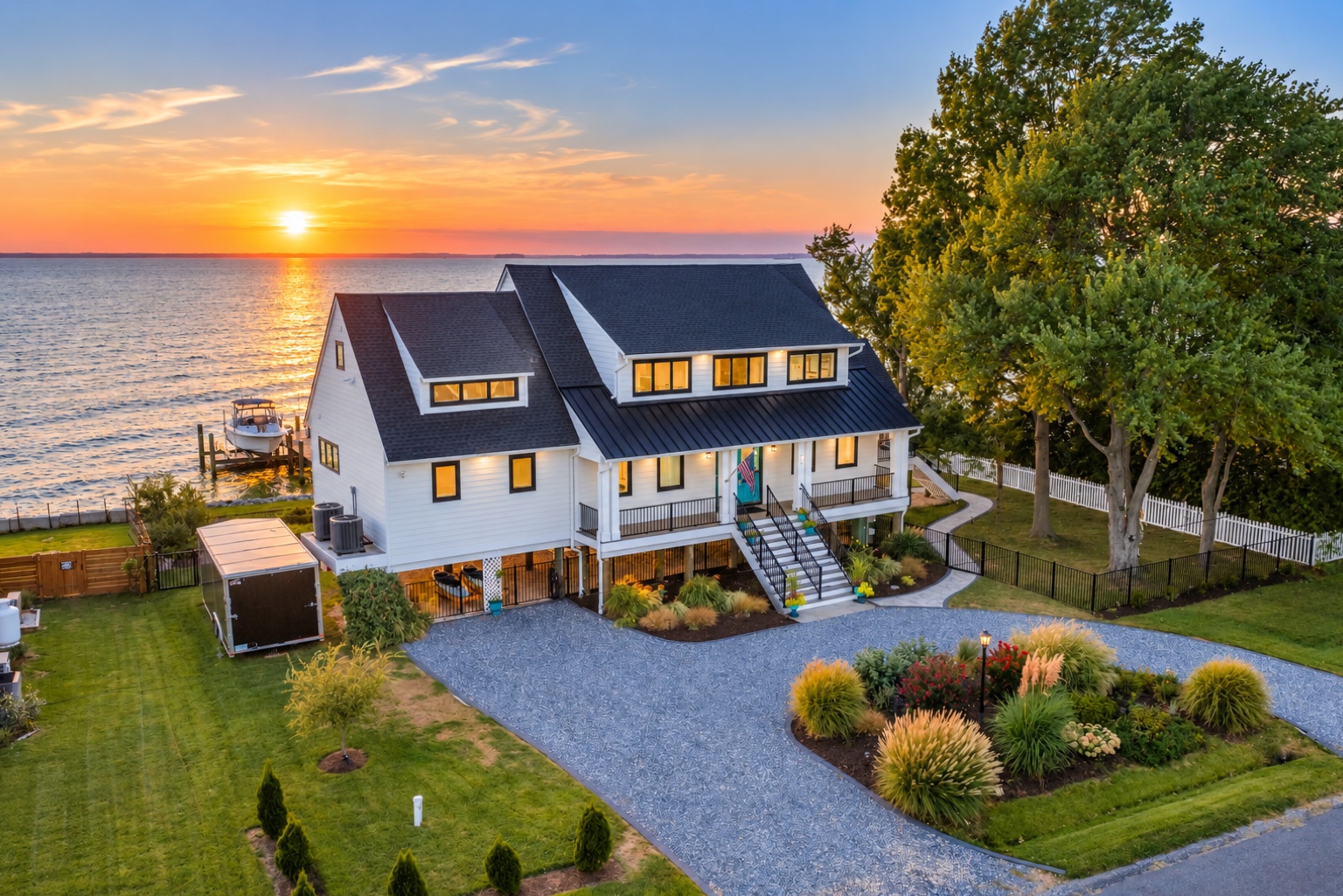 Large house with a dark roof, white walls, and multiple windows, situated by the water at sunset, surrounded by trees and a garden.