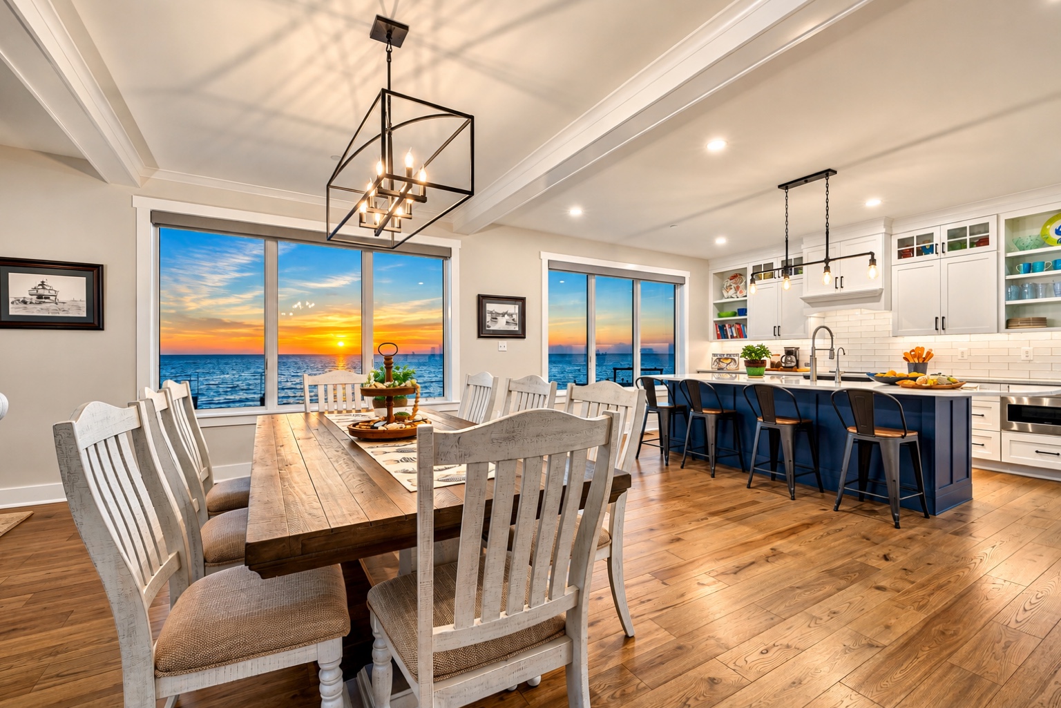 Open dining and kitchen area with large windows showing a sunset over water, wooden floor, and modern decor.