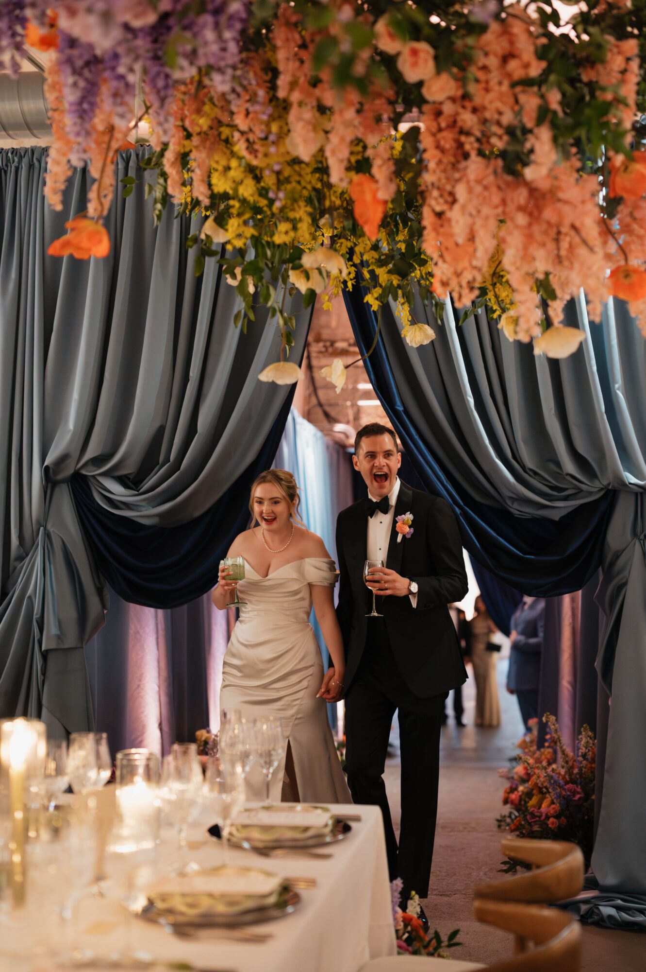 Bride and groom walking through decorated archway with hanging flowers, wedding reception table in foreground, elegant curtains behind, indoor setting.
