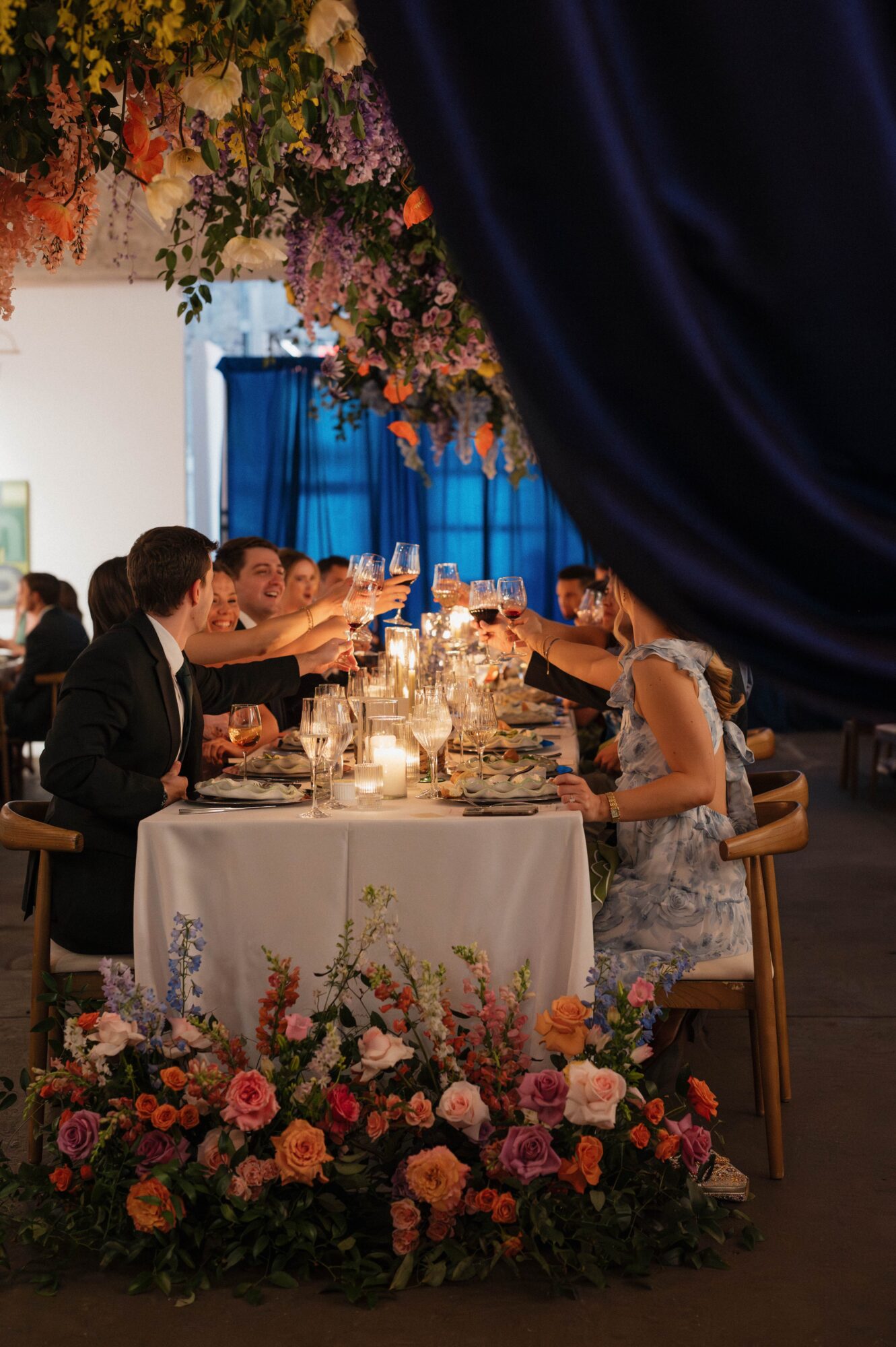 People seated at a decorated table with floral arrangements, dining in a dimly lit setting with blue curtains and hanging flowers.