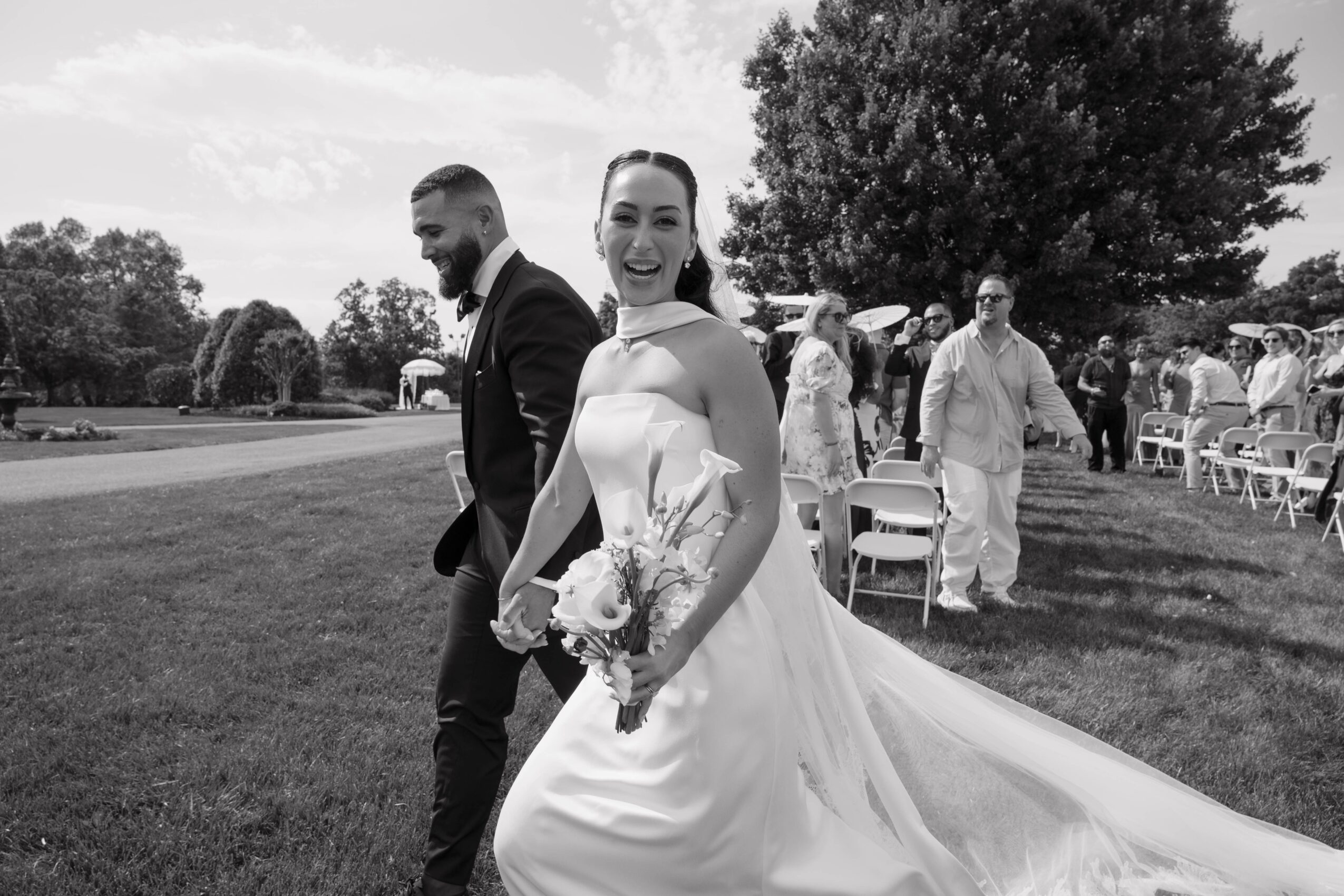 Bride in wedding dress holding bouquet, smiling, outdoors with guests and trees in background.