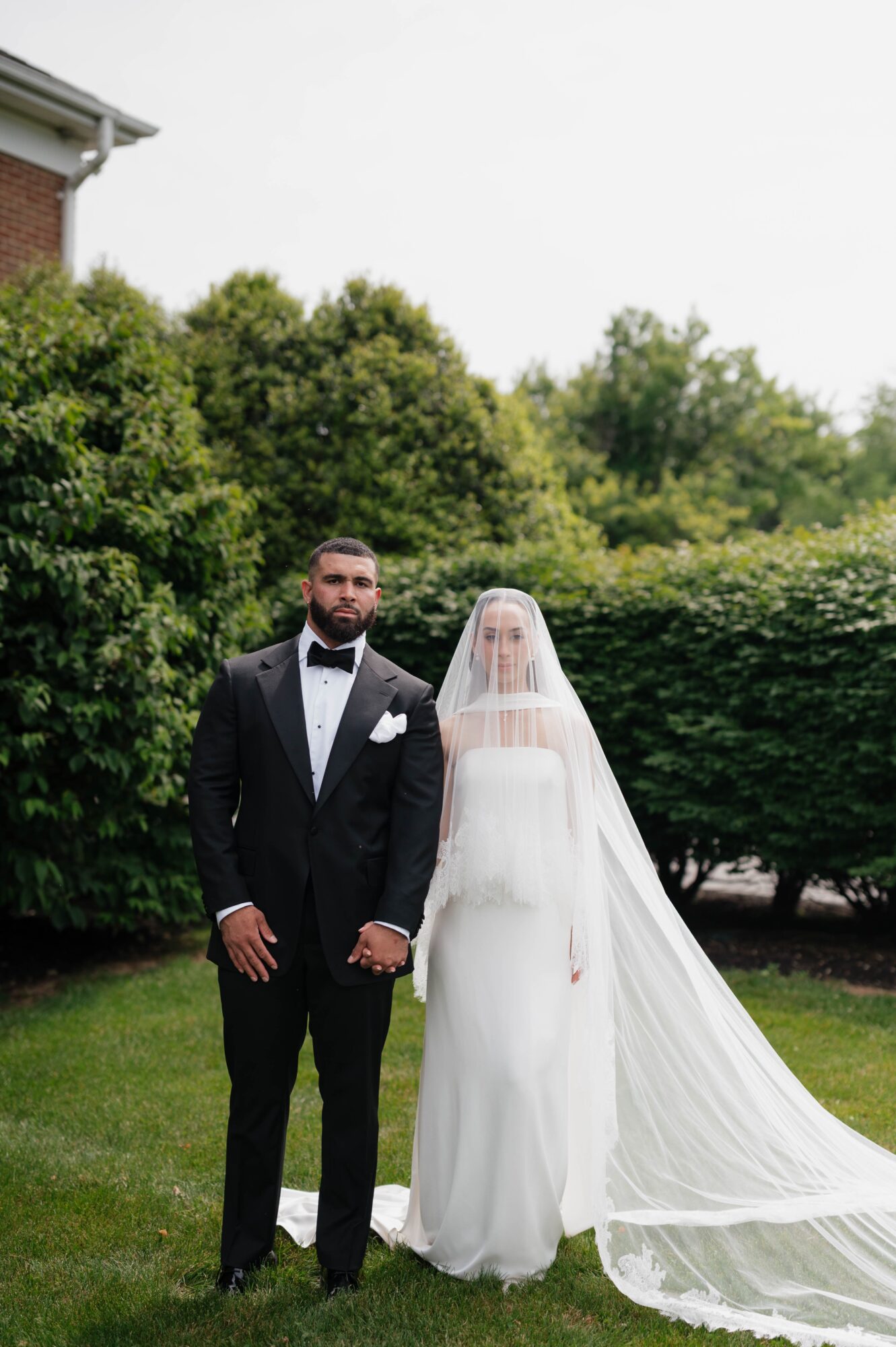 A bride and groom stand outdoors on grass, with trees and bushes behind them, dressed in wedding attire.