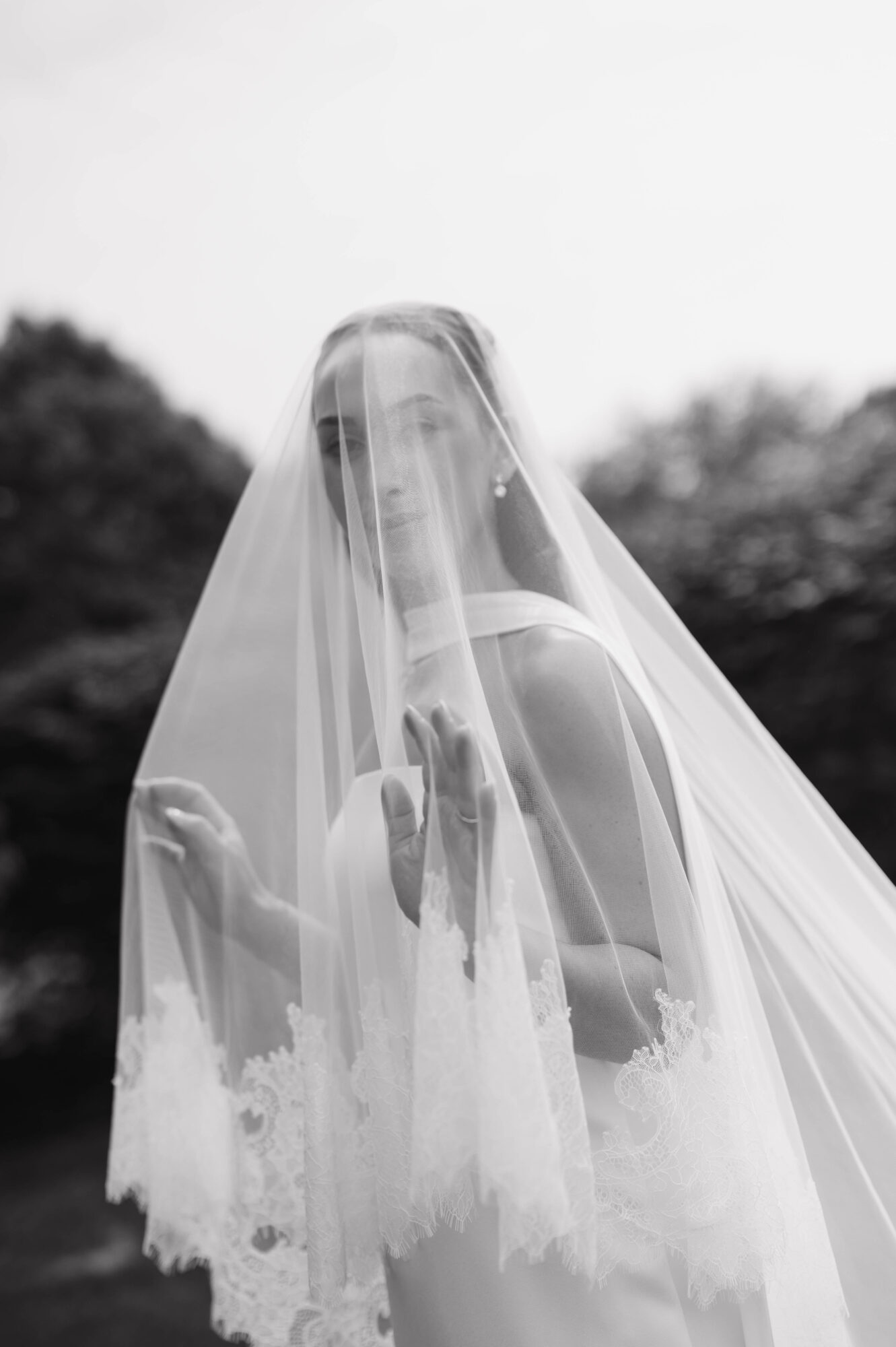 Person with a veil covering their face and shoulders, outdoors with trees in the background, black and white photo.