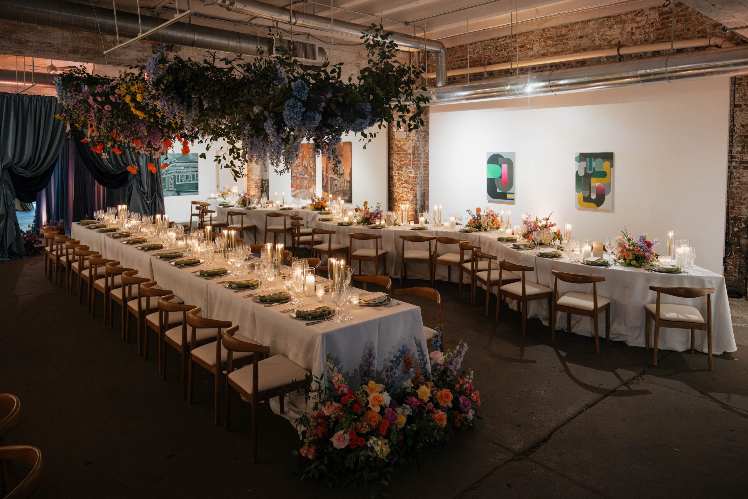 Long banquet table set with candles, flowers, and place settings in a decorated event space with hanging greenery and artwork on the wall.