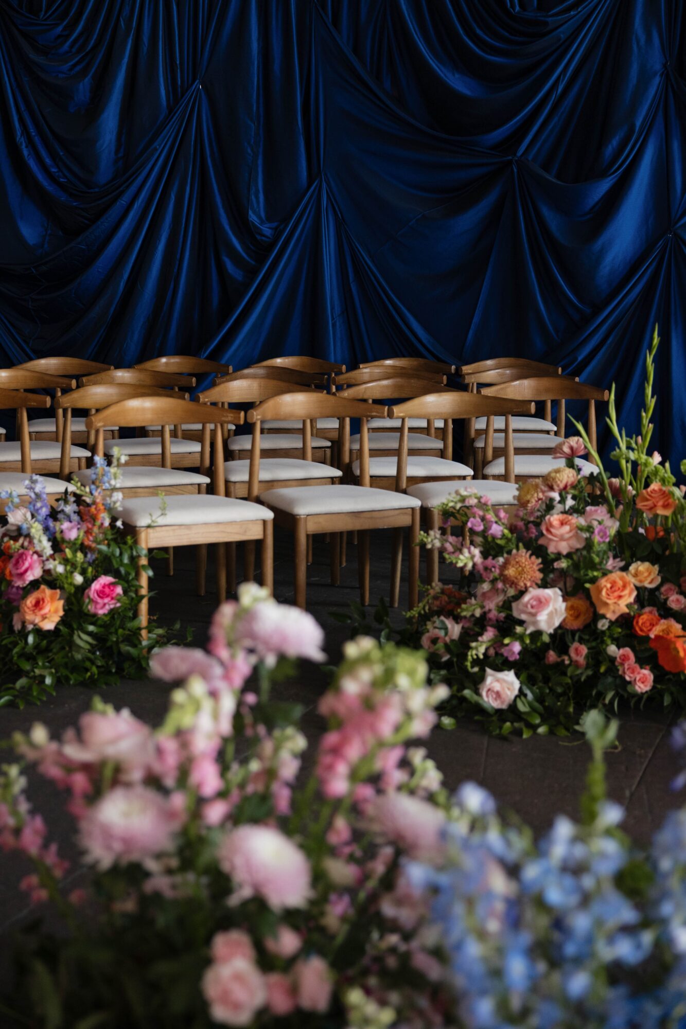 Rows of wooden chairs with white cushions in front of a dark blue curtain, surrounded by colorful flower arrangements.