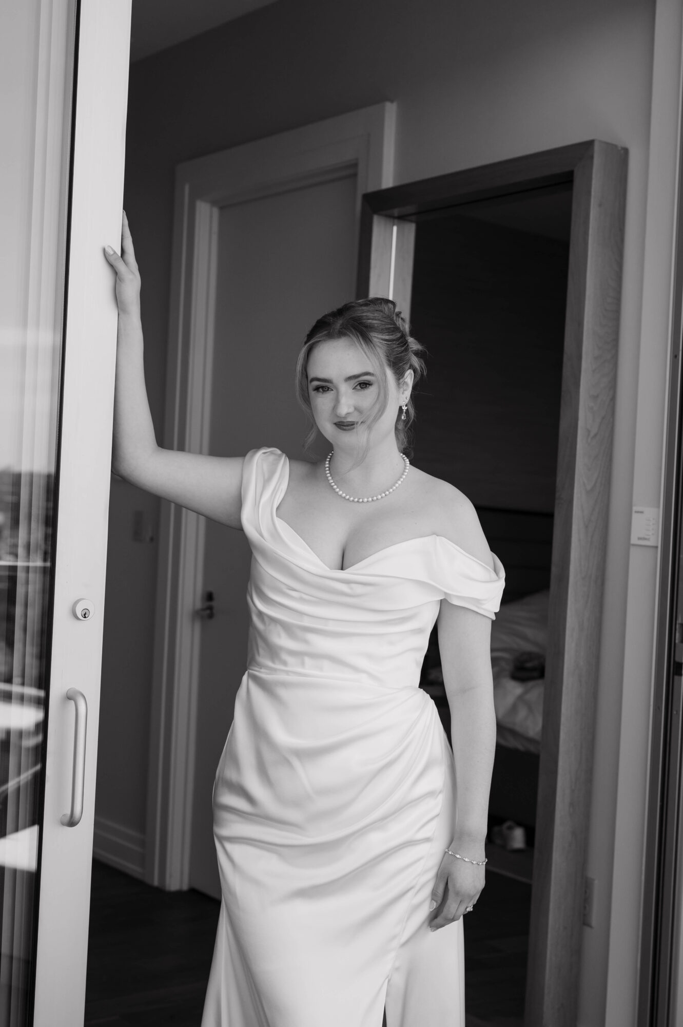 Young woman in a white dress standing in doorway, holding door frame, smiling, with a room visible behind her.