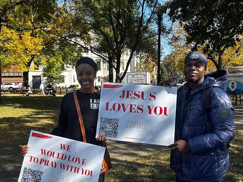 Two people holding signs in a park with trees and sunlight, promoting love and faith, smiling at the camera.