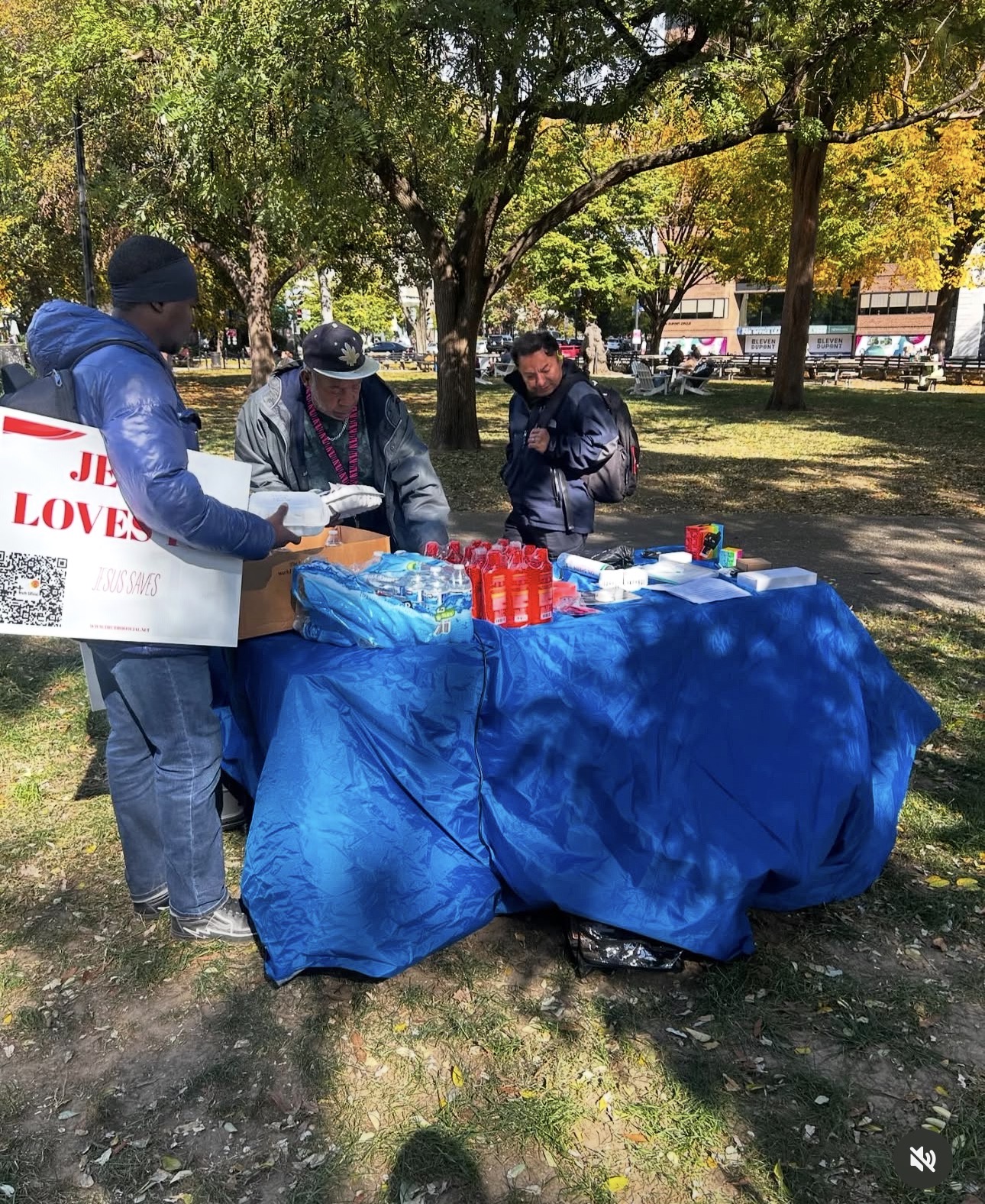 Three people gather around a table covered with a blue cloth in a park with trees and sunlight.