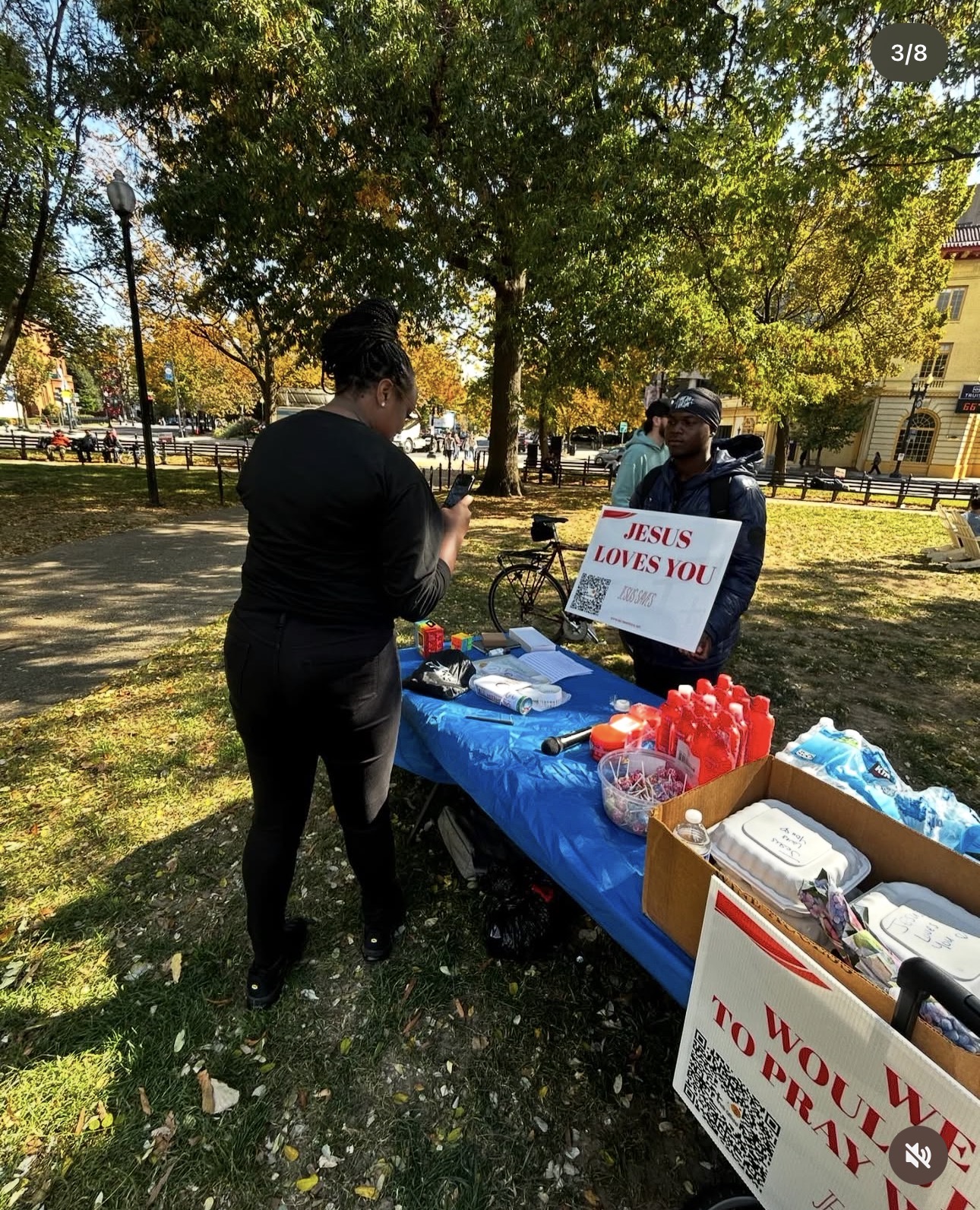 Two people at a table outdoors with a sign that reads 'JESUS LOVES YOU,' surrounded by trees and fall foliage.