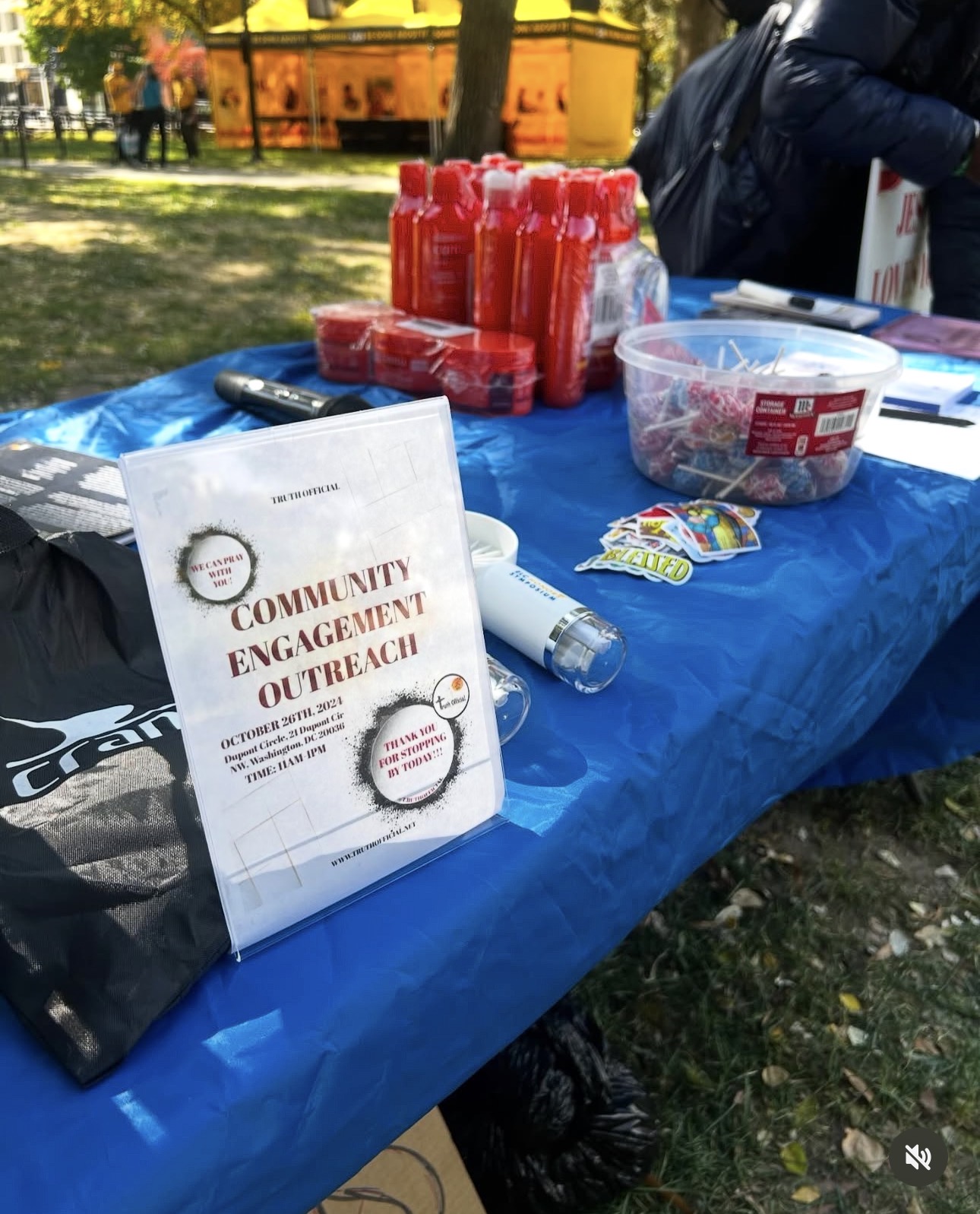 Table with red bottles, a bowl of candy, and a community engagement sign outdoors in a park.