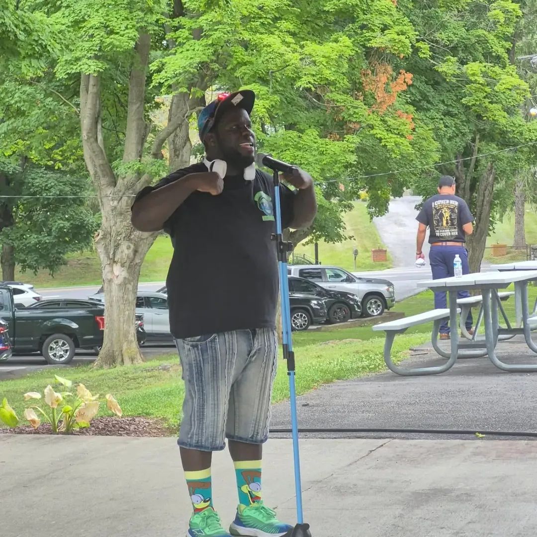 Person standing outdoors with trees and parked cars, holding a microphone, wearing a black shirt, striped shorts, and colorful socks.