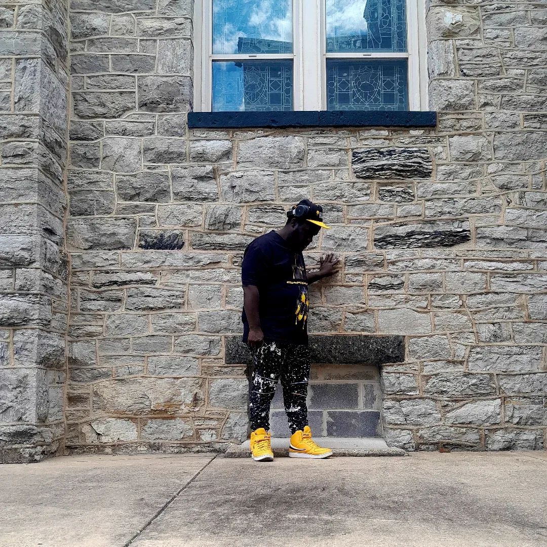 Person standing against a stone wall, looking at a phone, wearing black clothing and yellow shoes, near a window.