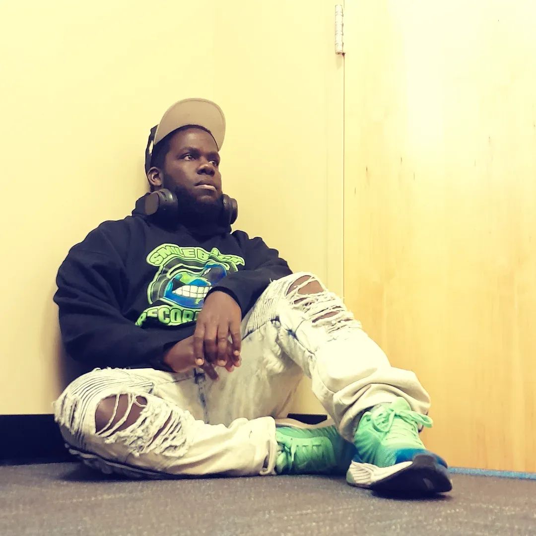 Young person sitting on the floor against a yellow wall, wearing a cap, headphones, black hoodie, ripped jeans, and sneakers.