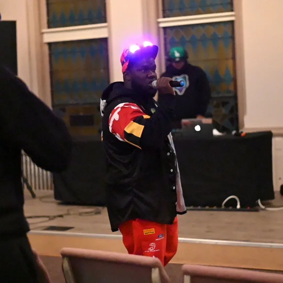 Person holding microphone, wearing colorful jacket and red pants, standing indoors with windows and DJ setup in background.