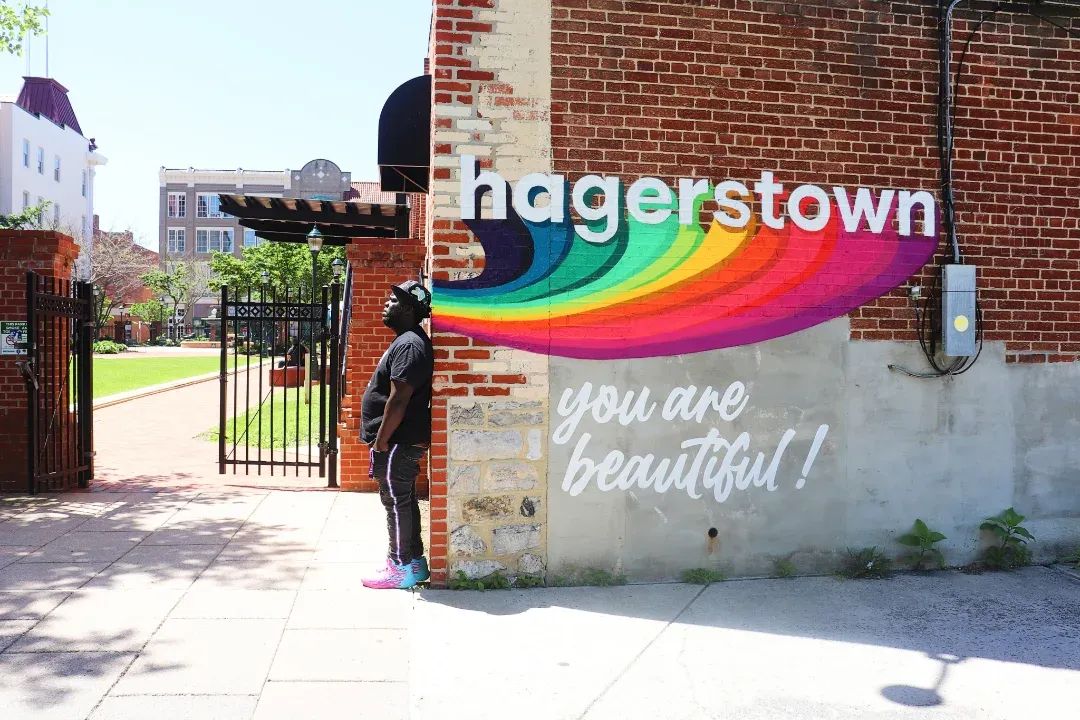 Person standing near a brick wall with colorful 'nagerstown' sign and white text 'you are beautiful!', sidewalk and gate visible.
