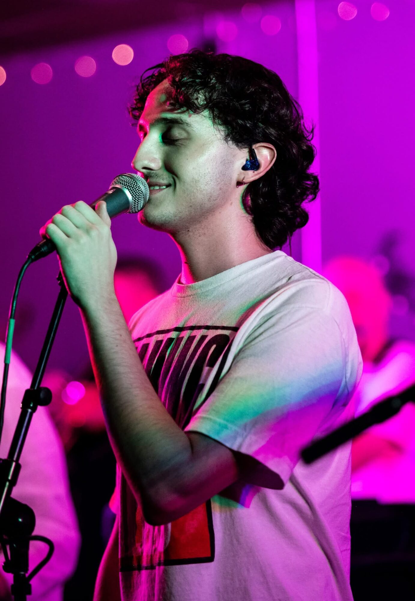 Young person singing into a microphone with eyes closed, wearing a white T-shirt, in a colorful stage setting.