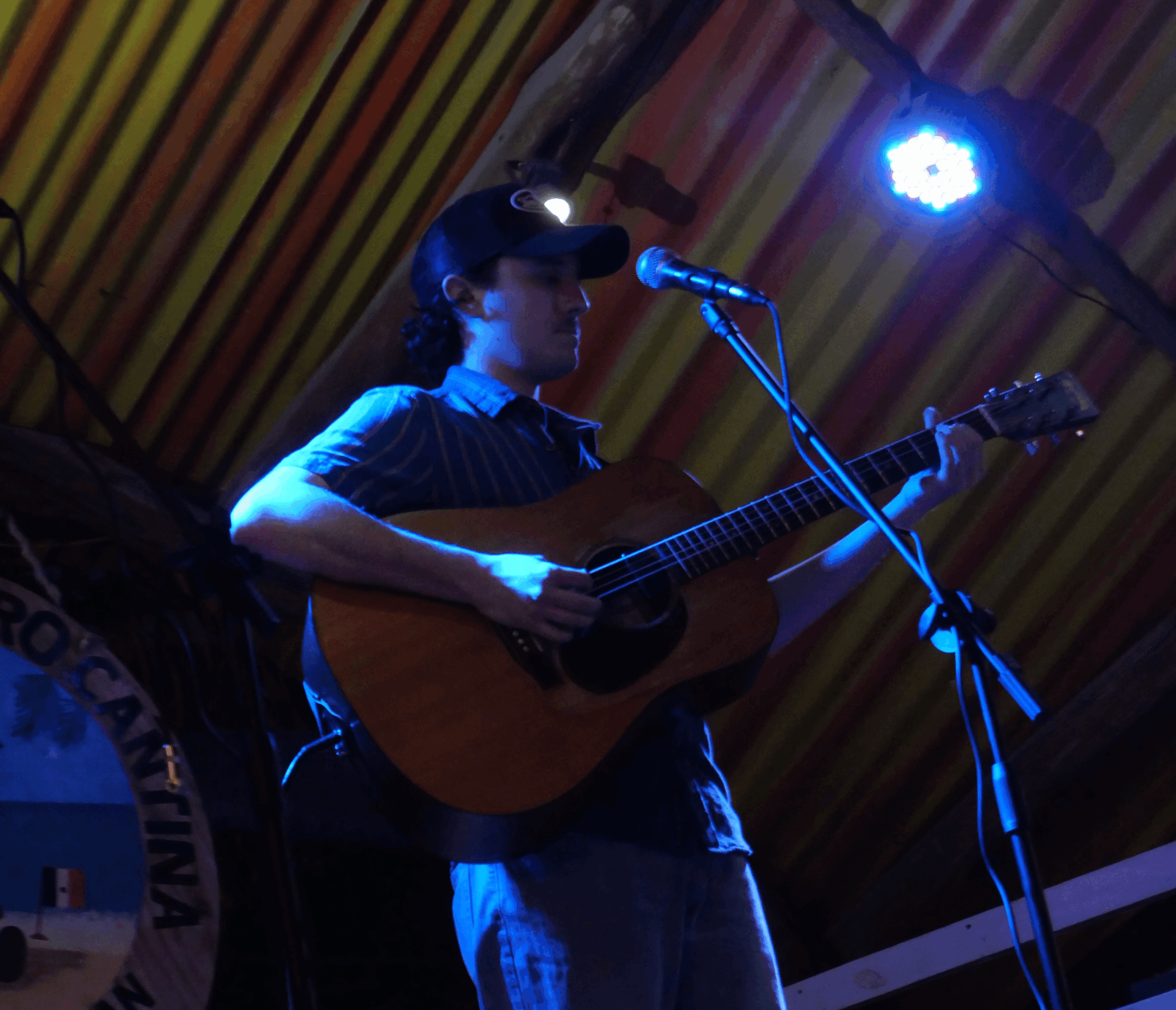 Person playing guitar and singing into microphone on stage with colorful striped background, illuminated by stage lights.