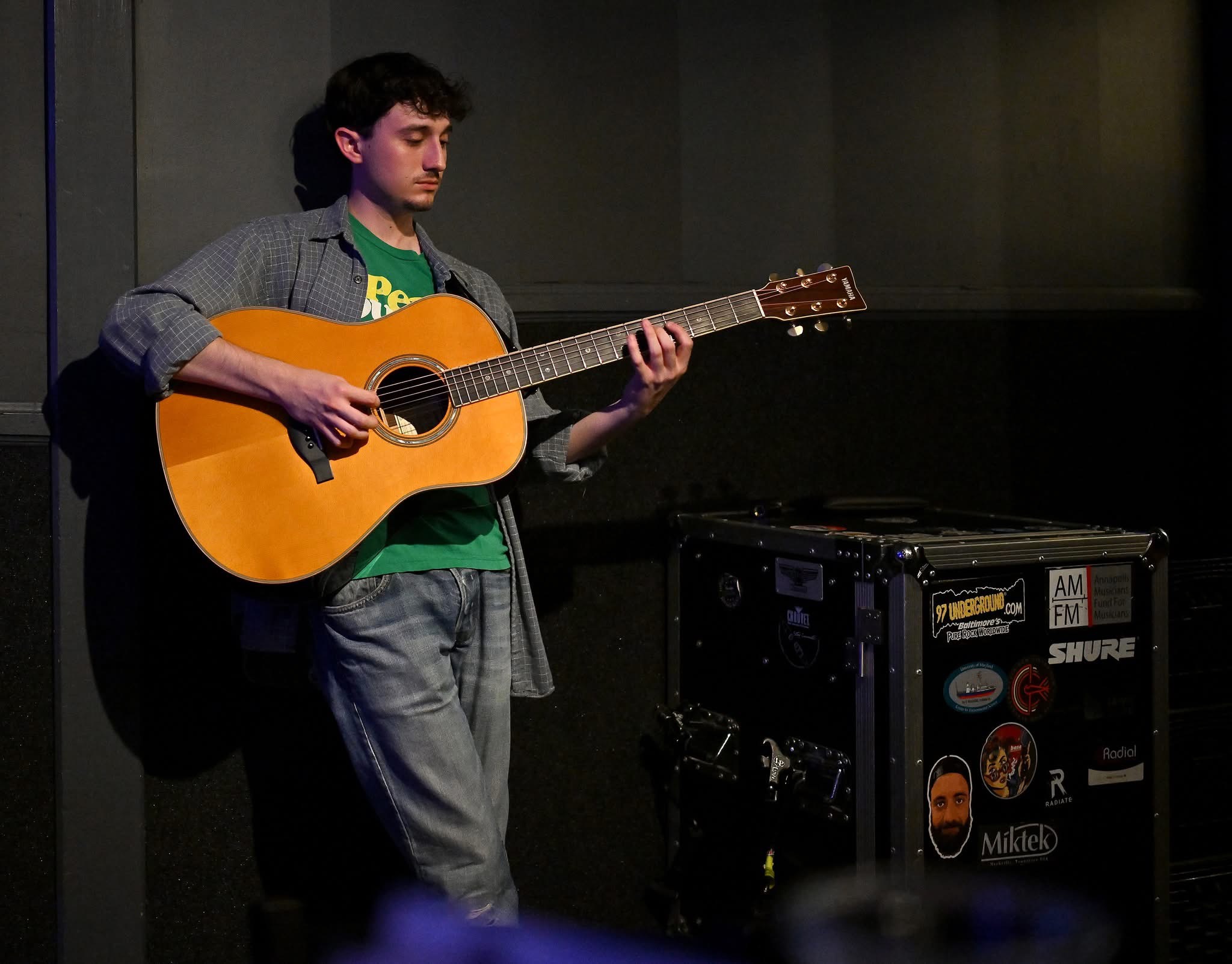 Young man playing acoustic guitar in a dimly lit room, standing next to a black equipment case with stickers.