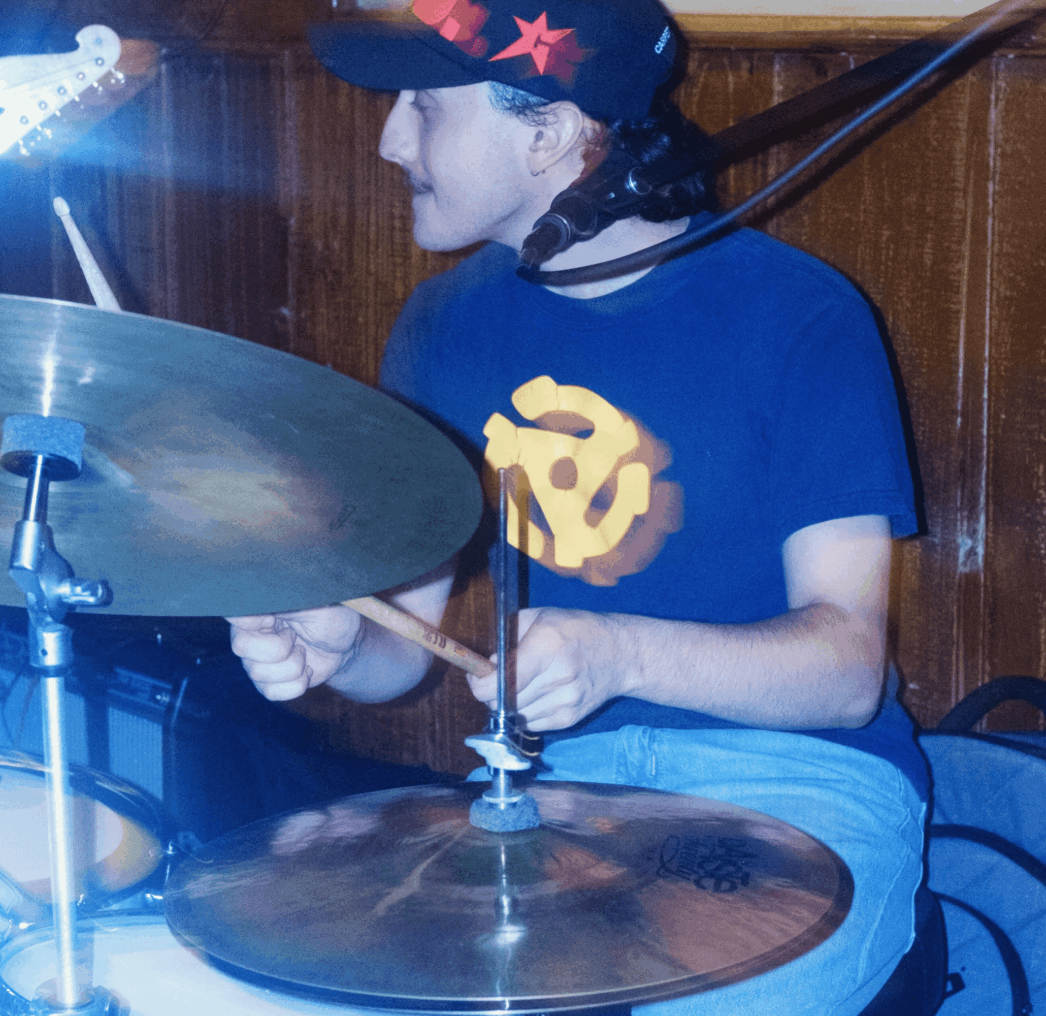 Young person playing drums, wearing a black cap with red crosses, blue T-shirt with a yellow design, and jeans, indoors.