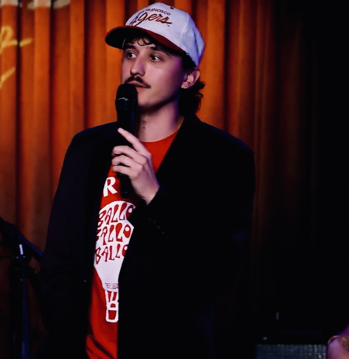 Person with dark hair, mustache, and beard, wearing a white cap, red shirt, and black jacket, holding a microphone.