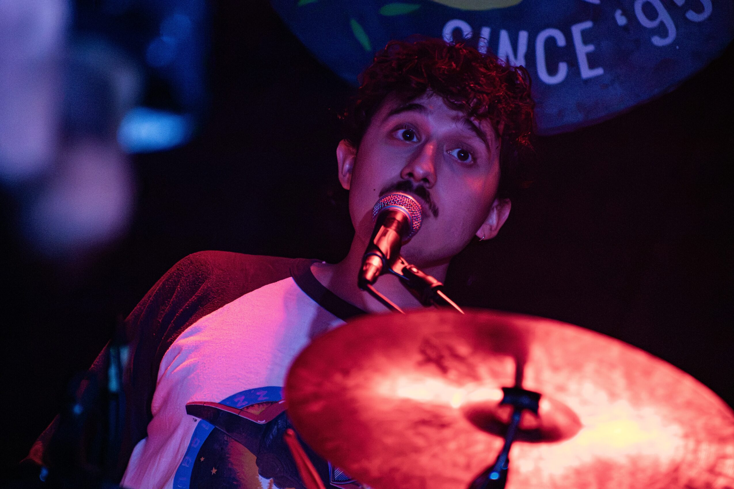 Young person singing into a microphone with a drum in front, stage lighting, and a dark background.