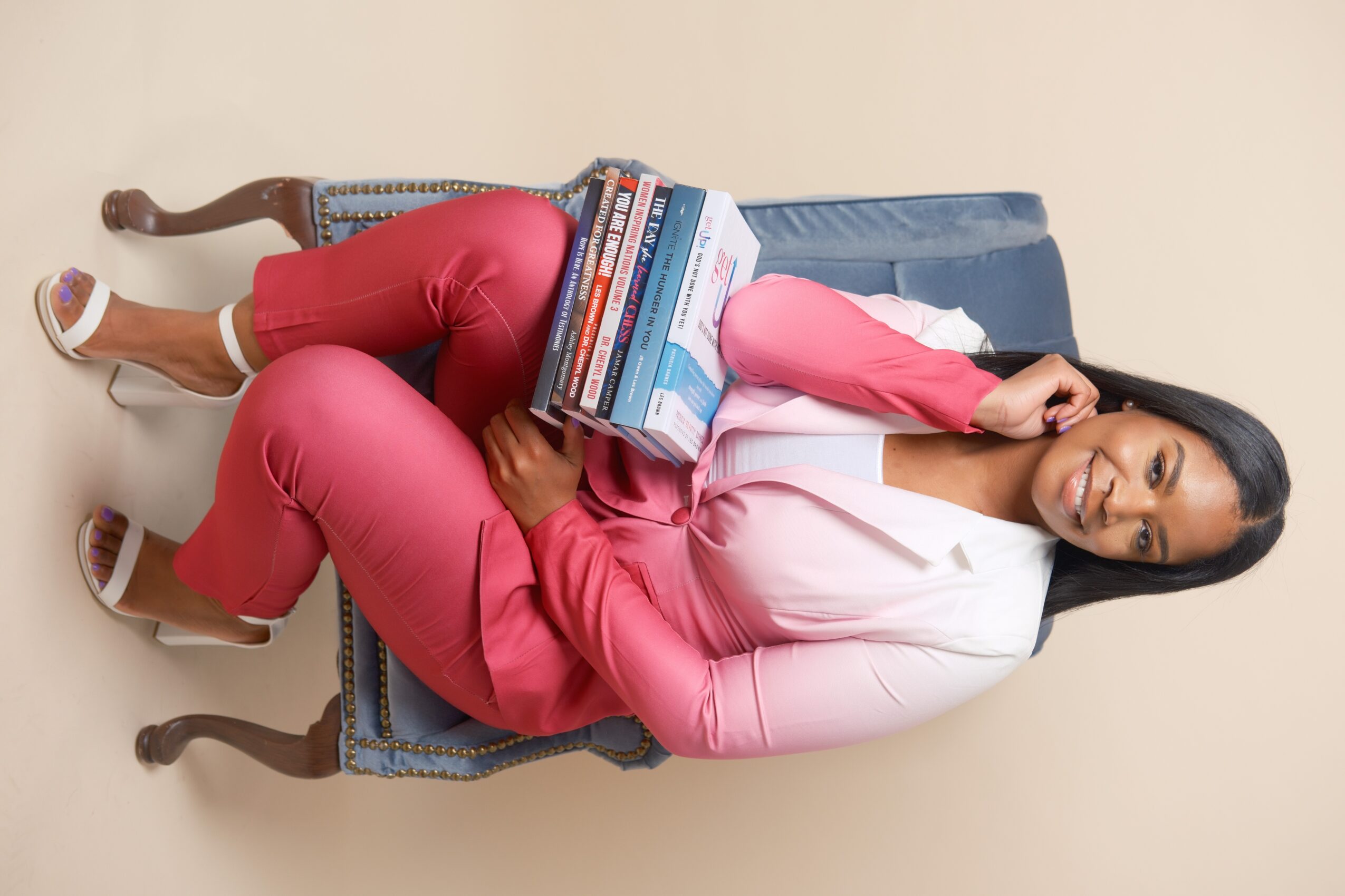 Woman in pink pants and blazer lying on a vintage chair, holding a stack of DVDs, against a plain beige background.