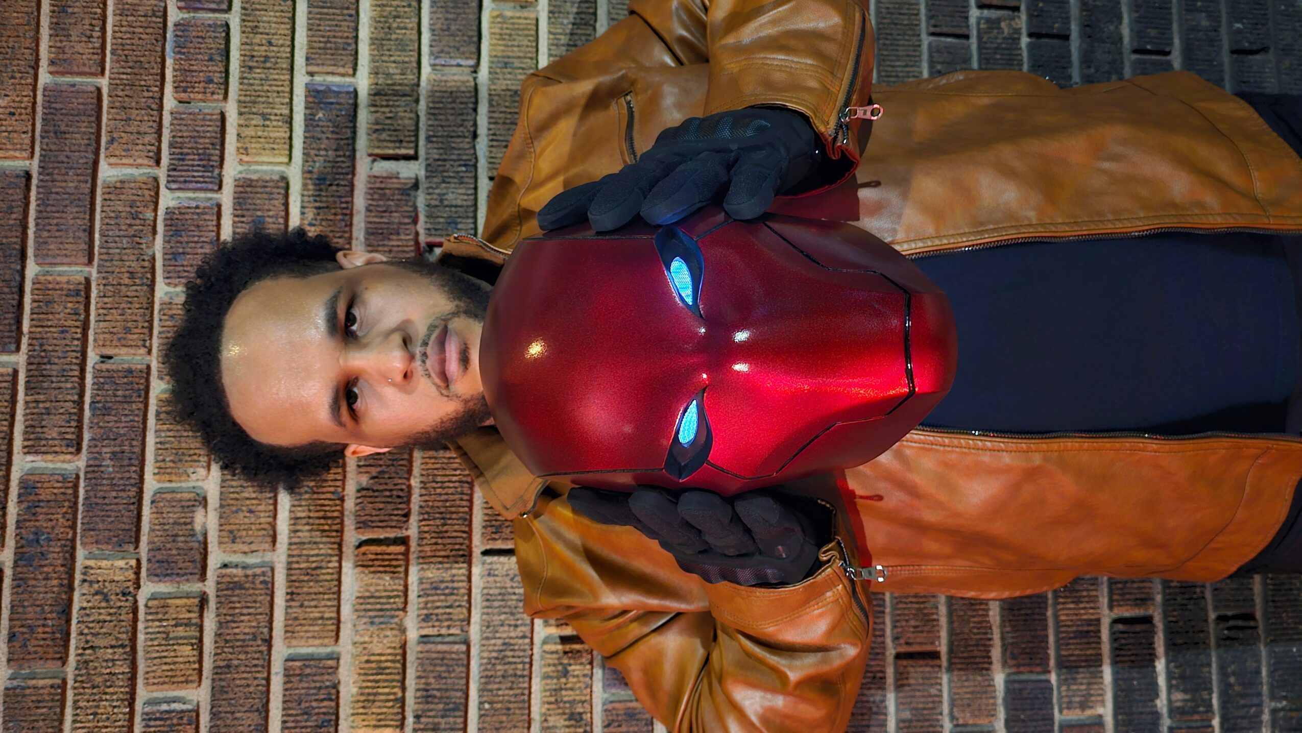 Person holding Iron Man mask with glowing eyes, standing against a brick wall, wearing a brown jacket and black gloves.