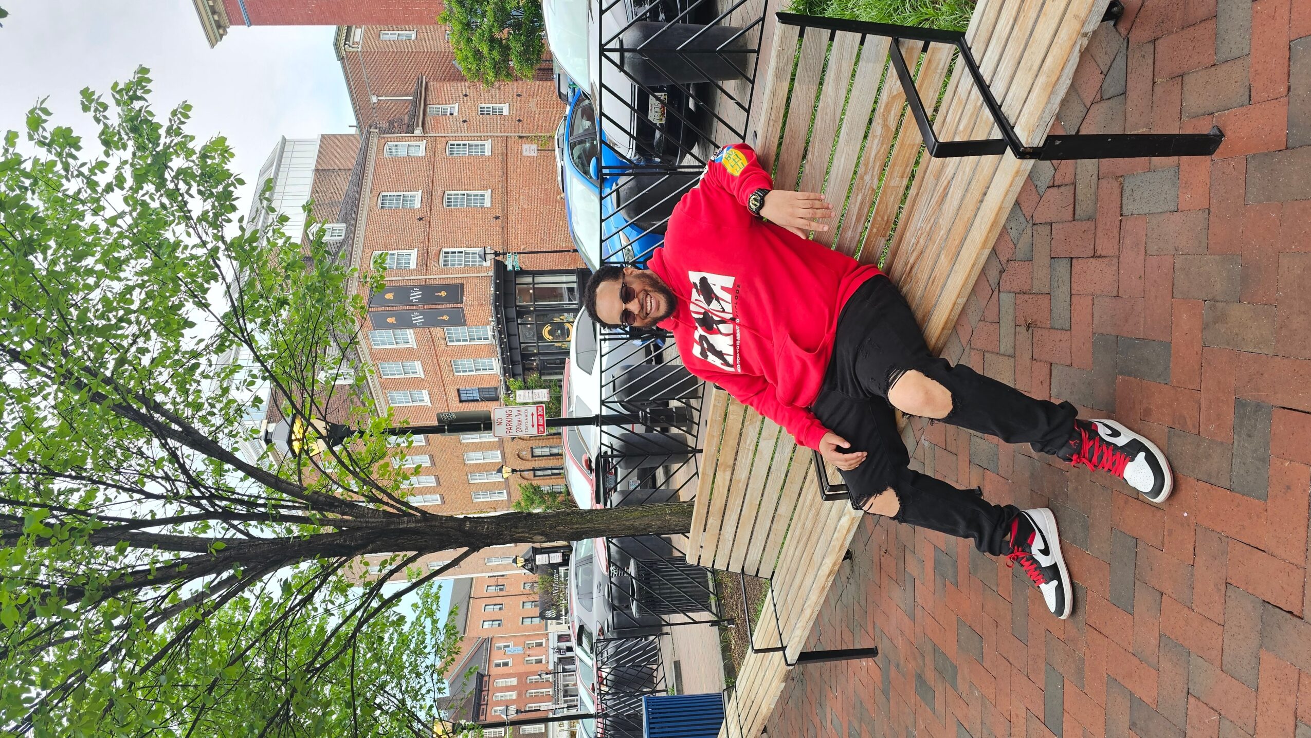 Person sitting on a bench outdoors, wearing a red hoodie, black pants, and sneakers, with trees and buildings in background.